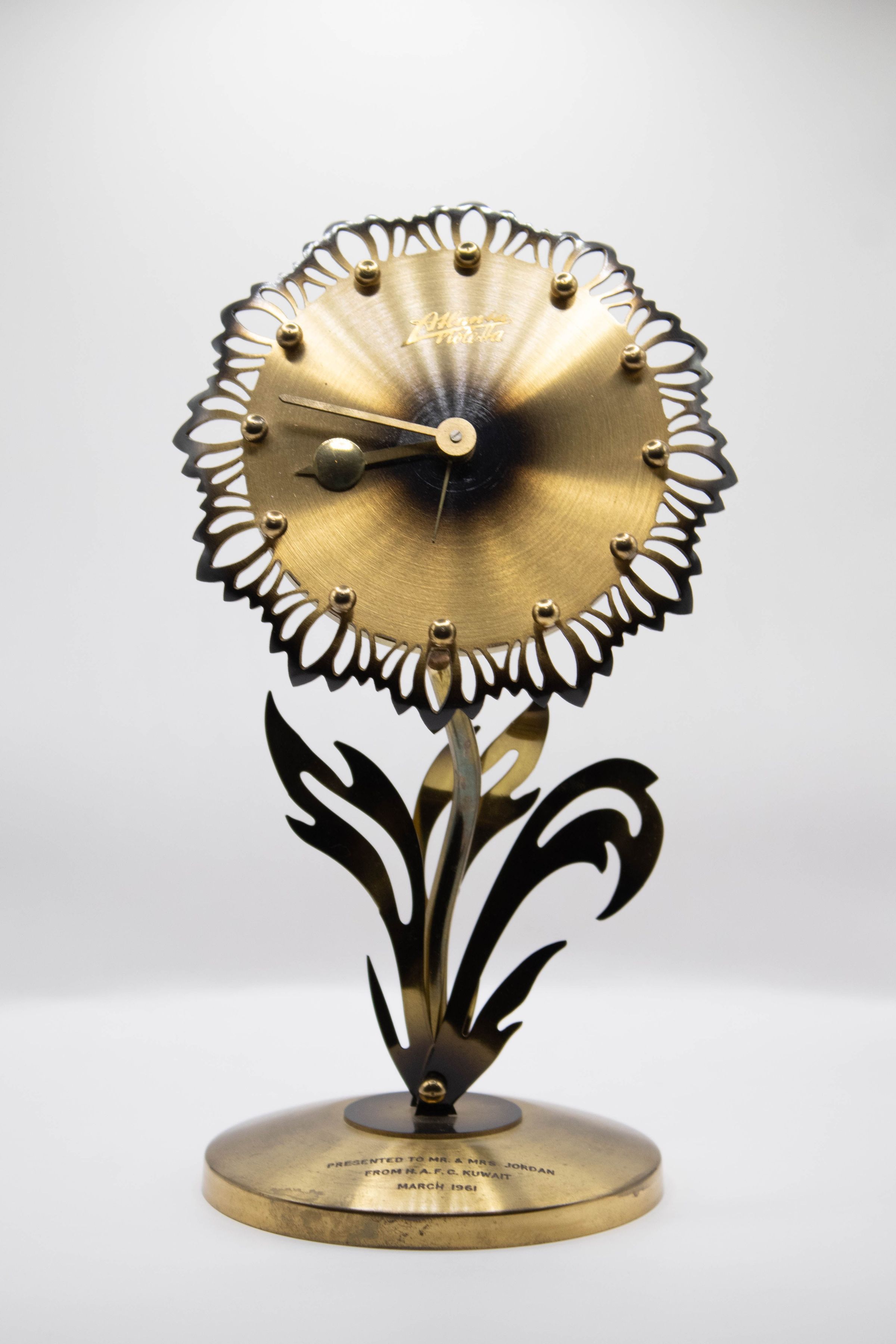 Brass Flower Clock: Image of the Brass Flower Clock. The clock is on a brass stem with leaves flaring out from the base. The brass leaves are darkened brass. The middle of the clock is a blackened brass with the second, minute, and hour hand radiating out from the middle. Rather than hours on the clock there are small brass balls in its place. There are darkened petals that line the outside of the clock. The name of the original maker, "Atlanta Violetta" is above the center of the clock.