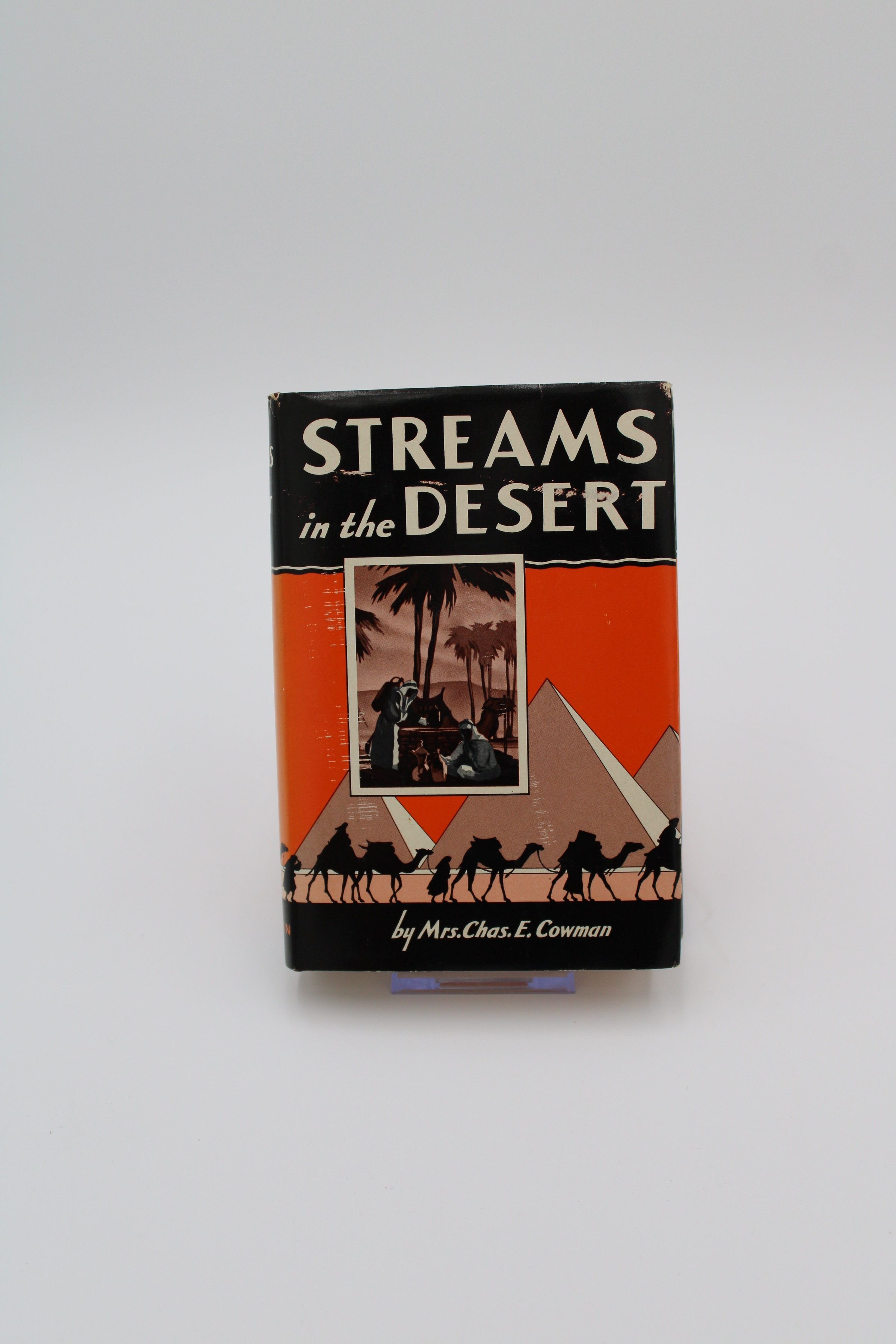 Front Cover: The front cover of “Streams in the Desert” features a depiction of a party of clothed travelers and their camels walking around the Egyptian Pyramids. The sky is orange while the sand is peach-colored. Atop the pyramid depiction is a rectangular box which features a scene of two Arabic men collecting water from a well.