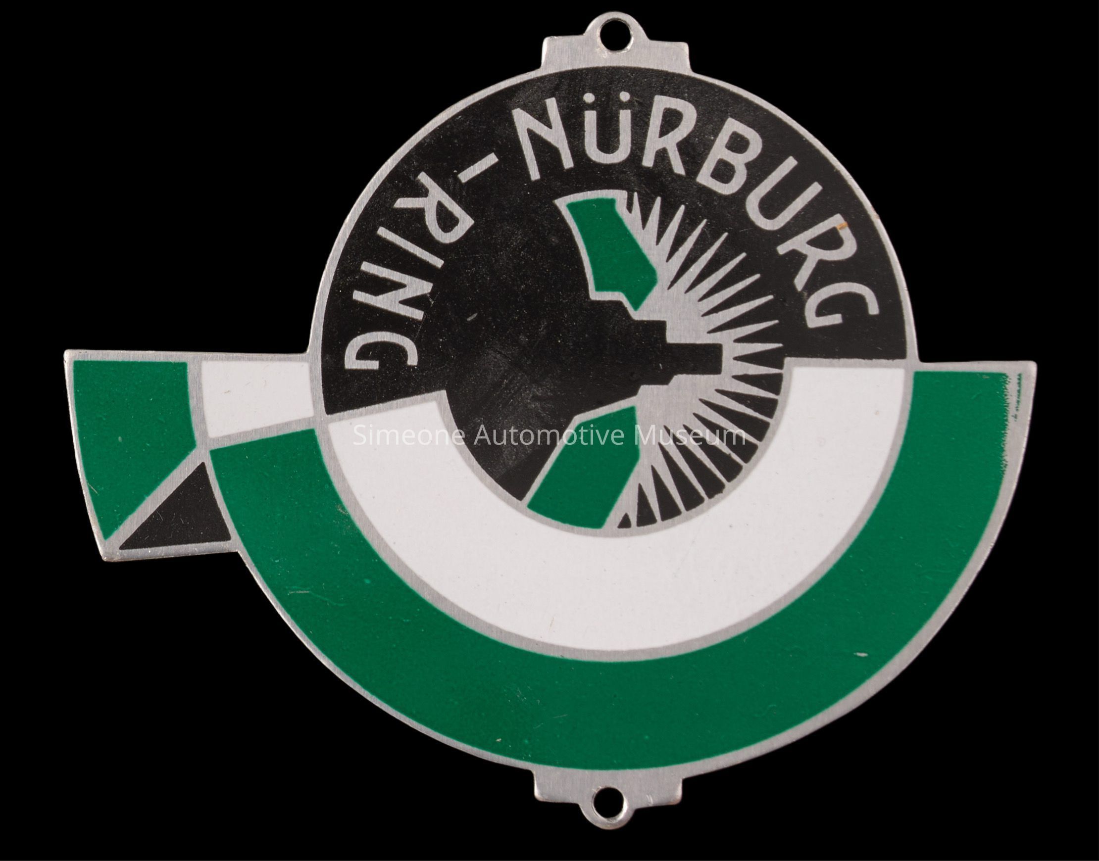 The image depicts a circular emblem featuring the name "NÜRBURGRING" written across the top in bold, white letters against a black background, while the lower portion of the circle is divided into a green and white geometric pattern, possibly representing a stylized racetrack or abstract design element; the black-and-white section includes a starburst effect, adding dynamic flair, while the entire emblem has a metallic sheen, suggesting it may be made of metal or chrome, and there is a small hole