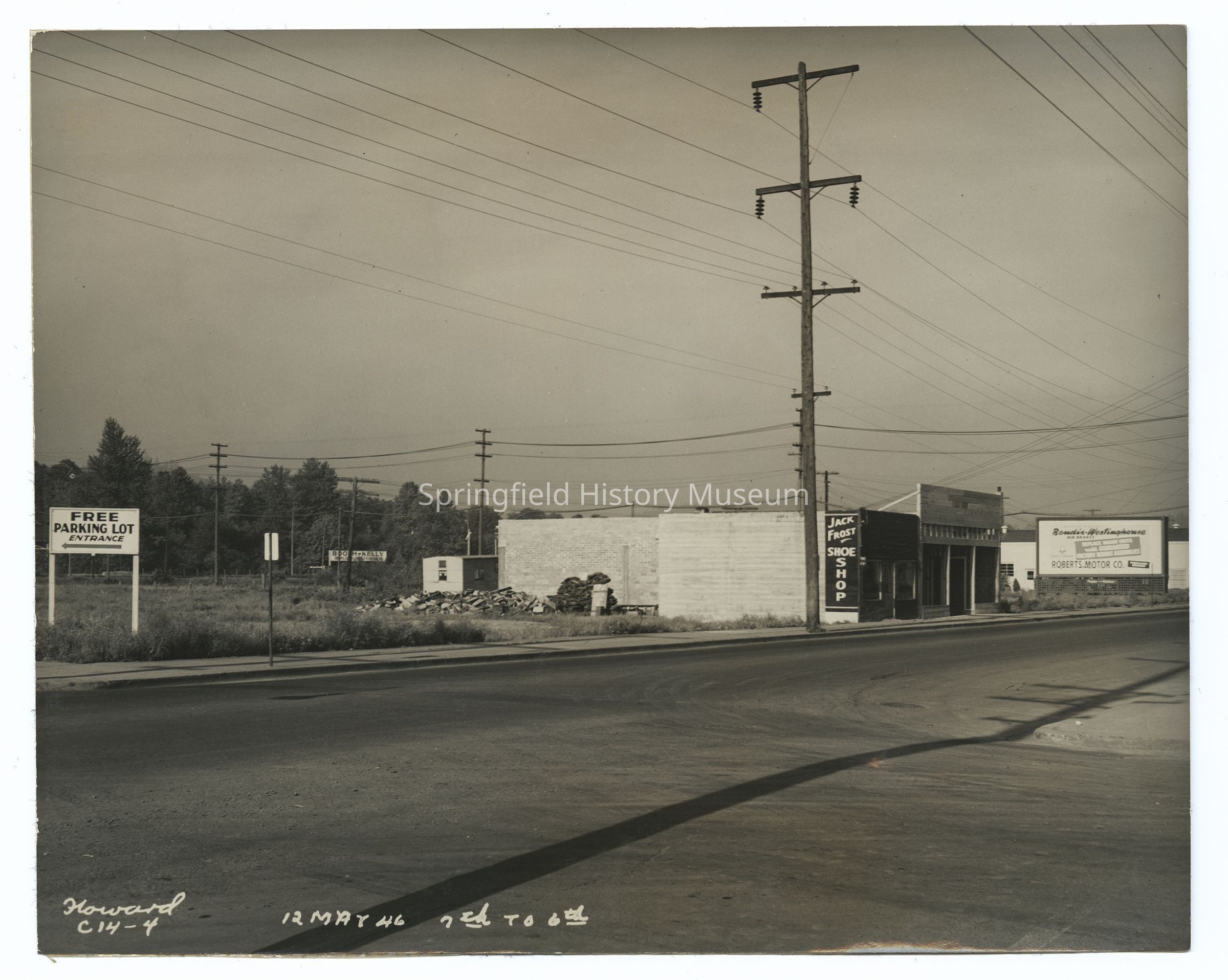 2021.005.049: The black and white photograph depicts Main Street with a large, empty parking lot on the left, marked by a "Free Parking Lot Entrance" sign. In the background, there are utility poles lined with power lines. A small building, labeled "Jack Frost Shoe Shop," sits along the road with its simple facade. Another sign advertises "Robert's Motor Co.," indicating a nearby business.