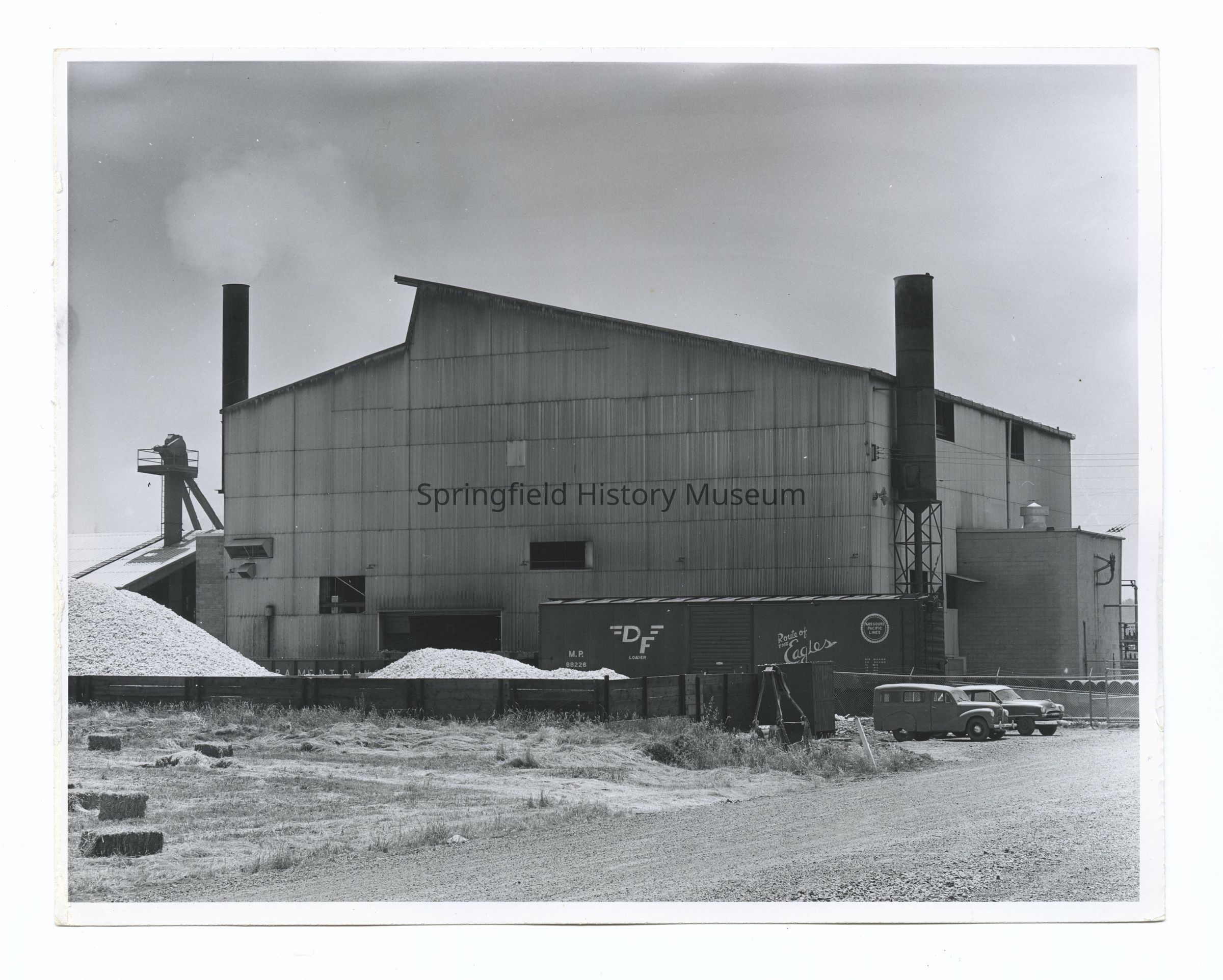 2021.005.005: The black-and-white image depicts a large industrial building with two tall smokestacks, releasing smoke into the sky. In front of the corrugated metal structure, there are piles of materials, protected by a fence, suggesting an outdoor storage area. A boxcar marked "DF" is on a track next to the building, hinting at rail access for transporting goods. Two vintage cars are parked on a gravel area, contrasting the industrial setting with elements of everyday life.