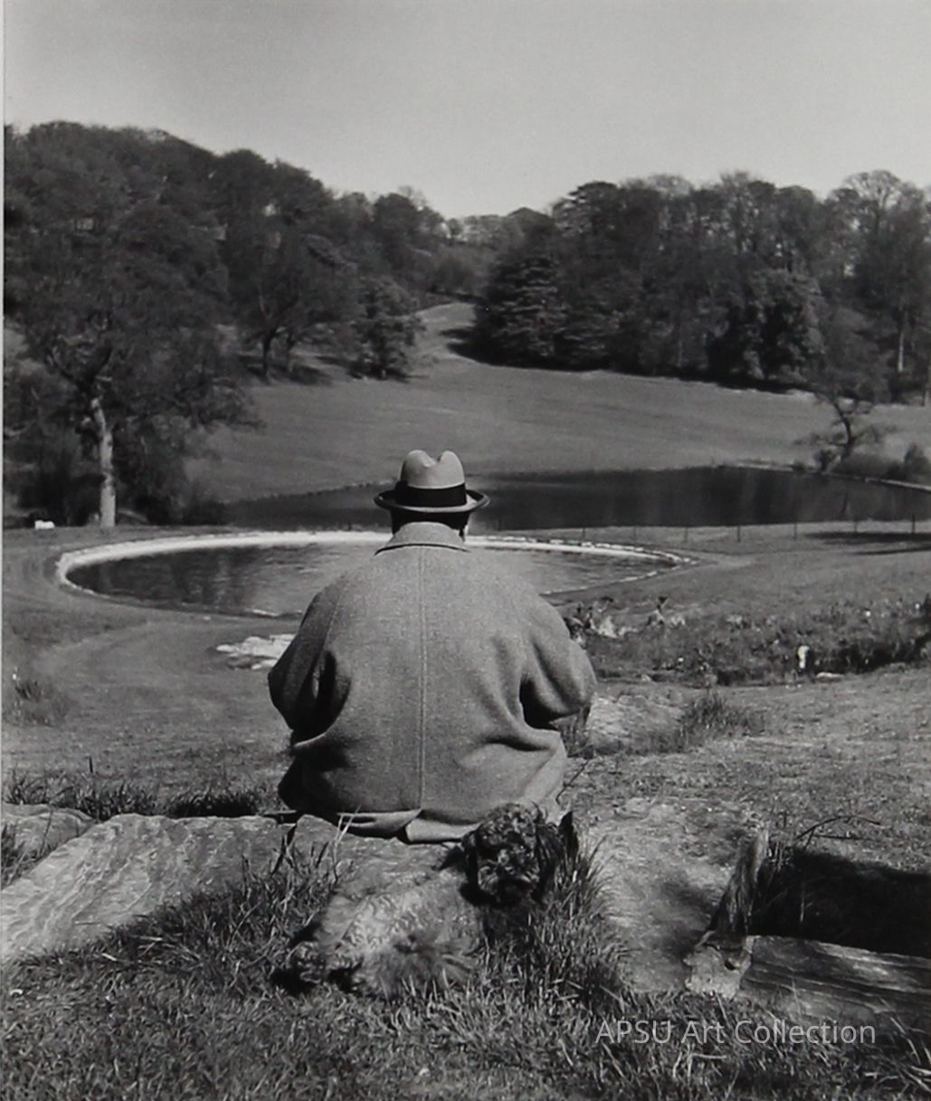This black and white photograph features a serene scene where a man in a hat and coat sits on a rock, overlooking a peaceful lake surrounded by lush trees. His relaxed posture and the tranquil natural environment evoke a sense of quiet contemplation and connection with nature. The curvature of the lake's shore and scattered light through the trees add depth and texture to this picturesque landscape, capturing a moment of solitude and reflection.