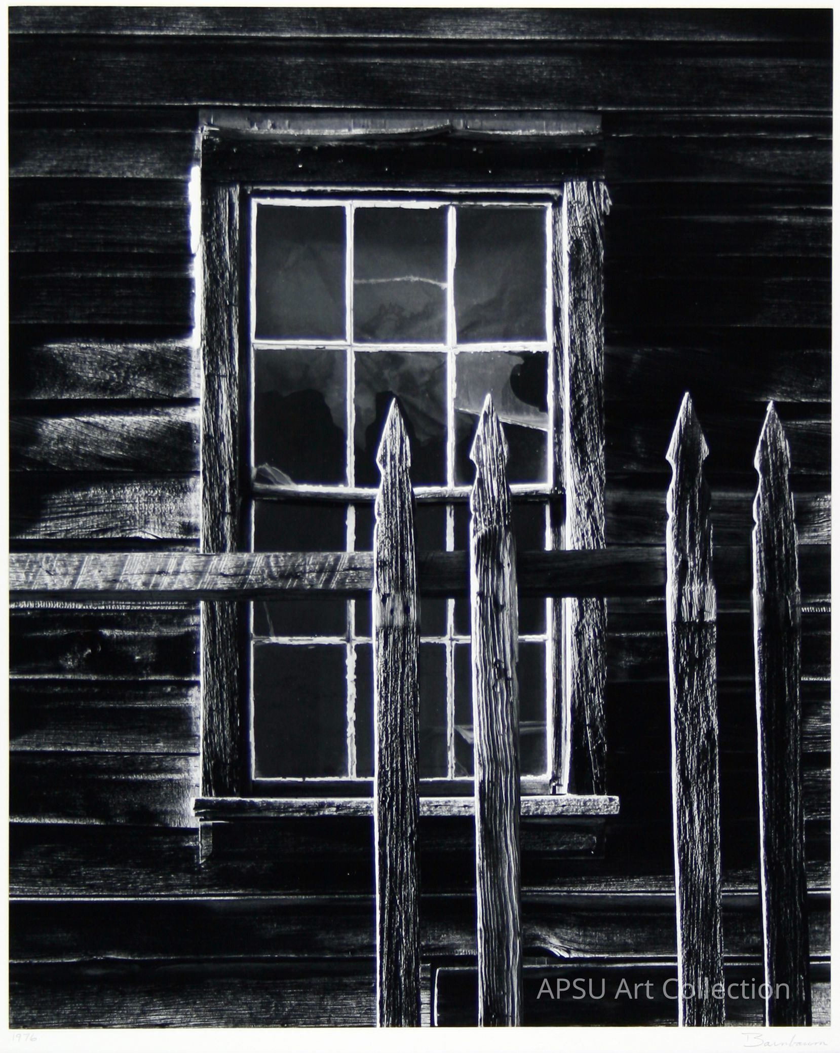This black and white photograph depicts a rustic wooden window on an old, weathered building, partially obstructed by a fence with pointed wooden pickets in the foreground, all bathed in dramatic lighting that highlights the texture and grain of the wood, creating a contrast between light and shadow, contributing to the nostalgic and somewhat eerie atmosphere of the composition.