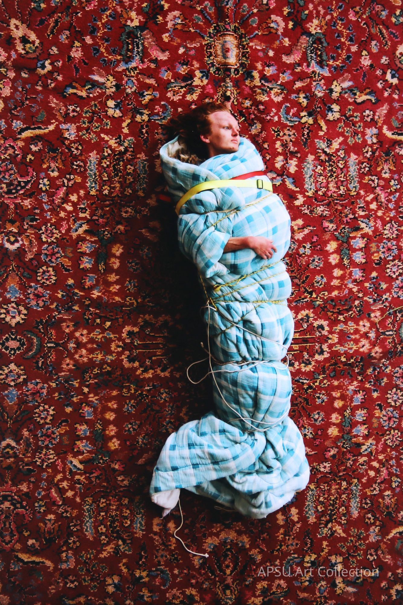 In this striking image, a person with long reddish-brown hair is bound in an exaggerated, cushioned swaddle of blue and white plaid fabric, secured with yellow rope, against a vibrant red and ornately patterned carpet backdrop, creating an intriguing and surreal visual contrast that suggests themes of confinement and comfort.