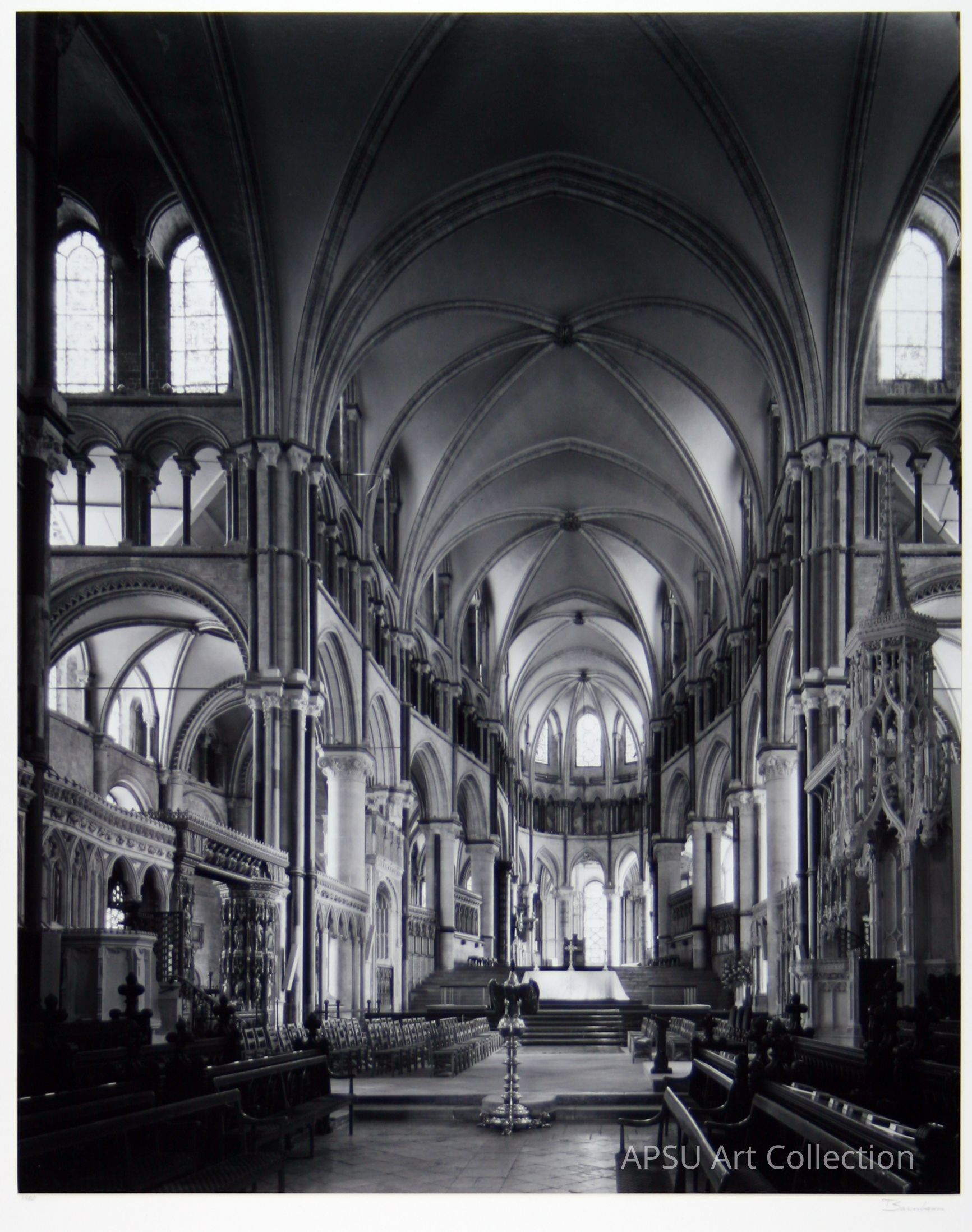This black and white photograph captures the stunning interior of a grand cathedral, characterized by its high, pointed arches and ribbed vaulting in the ceiling. The architectural style is reminiscent of Gothic design, with intricately carved columns and arched windows letting in light. The image beautifully showcases the symmetry and elegance of the structure, highlighting the detailed stonework, grand pillars, and the arrangement of benches leading towards the altar. The overall ambiance conveys a sense of reverence and historical grandeur.