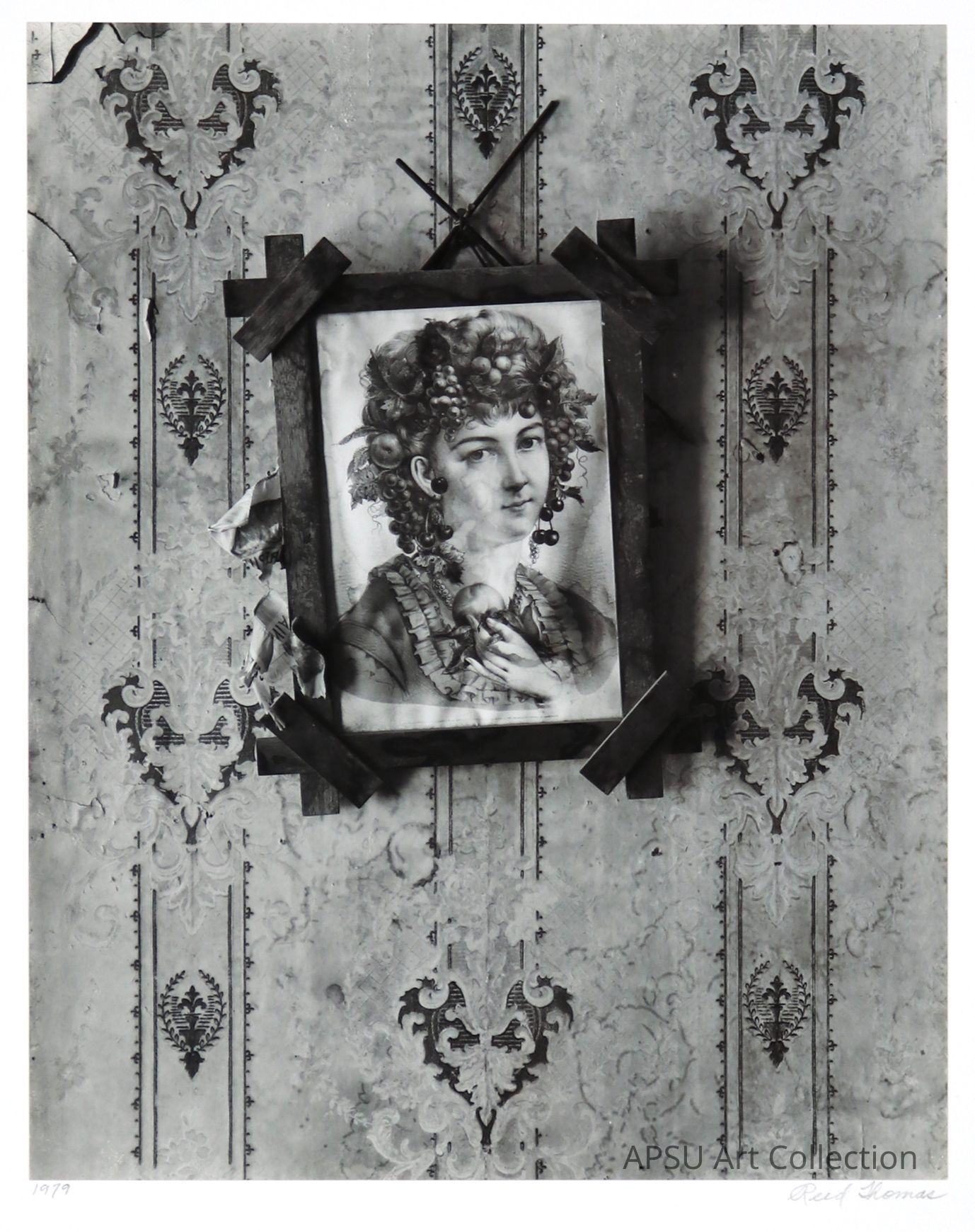 The image shows a black-and-white photograph of a circular frame containing a portrait of a woman with elaborate hair adorned with grapes and leaves, set against a textured backdrop of floral wallpaper. The frame is crossed by black tapes, creating a dramatic contrast with the elegant décor. The wall is decorated with intricate and symmetric patterns that add depth to the composition.