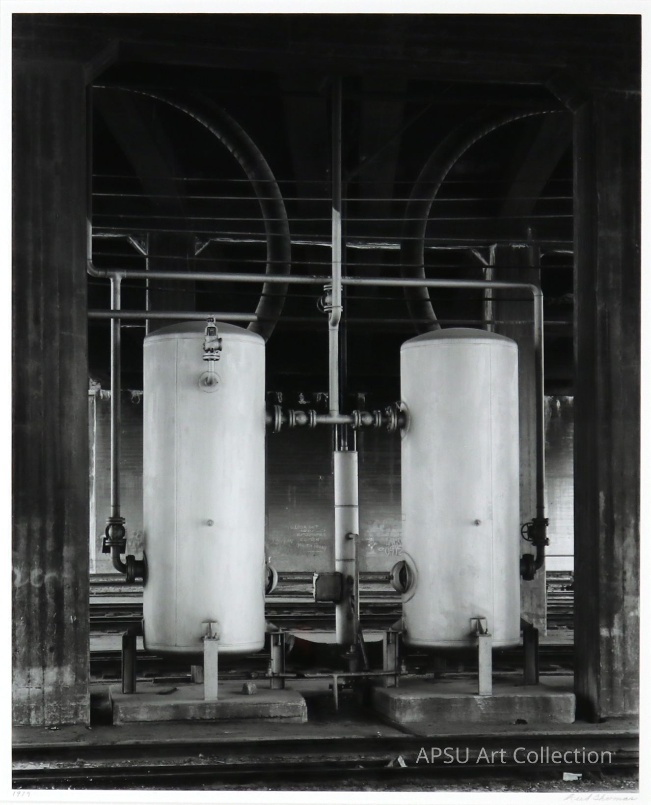 The image depicts a monochromatic industrial setting featuring two large vertical cylindrical tanks enclosed in a robust metallic framework with an array of pipes and valves attached, all housed within a dark, structured compartment under an arching network of additional piping, offering a detailed glimpse into a mechanical, possibly factory-related environment.