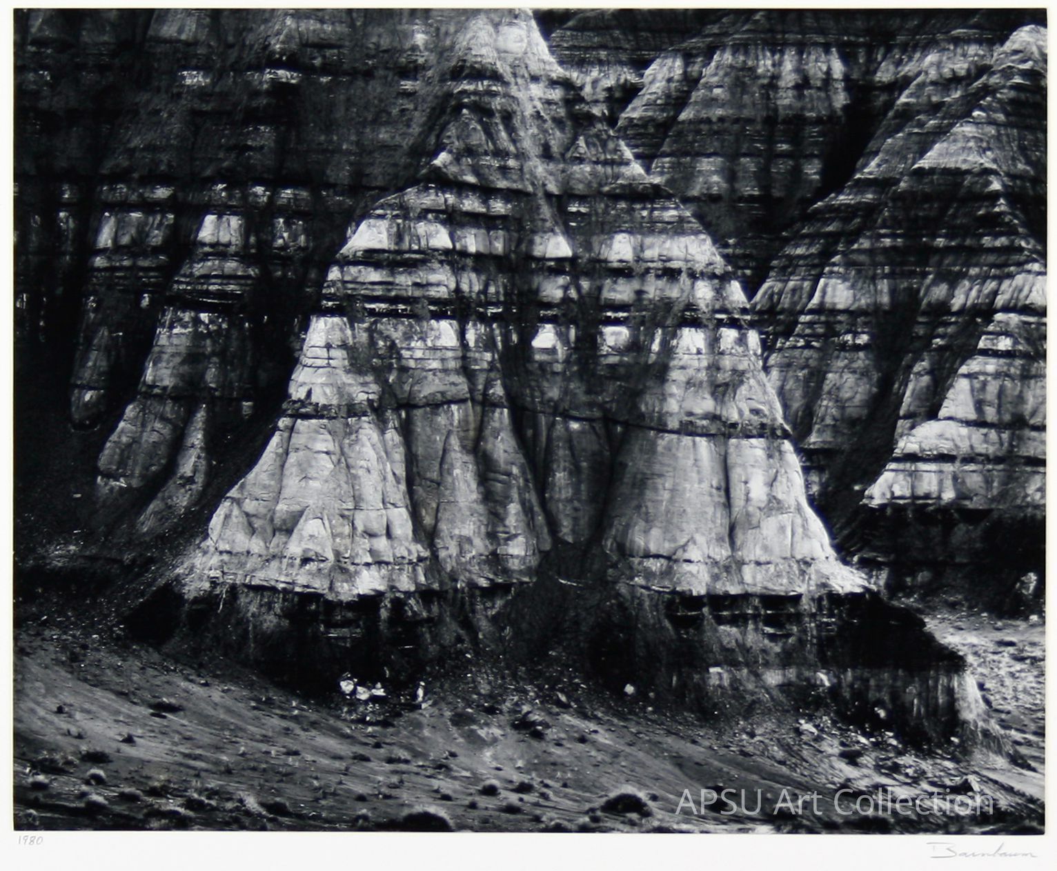 The image depicts a series of layered rock formations in a stark black and white photograph, where the erosion and stratification of the rocks are clearly visible, creating a visually striking contrast between the dark and light layers; the detailed texture suggests a geological formation shaped over countless millennia, encapsulating the rugged beauty of nature’s slow, relentless artistry. The bottom of the photo shows a scattered, rocky foreground, gradually blending into the monumental cliff faces that dominate the image.