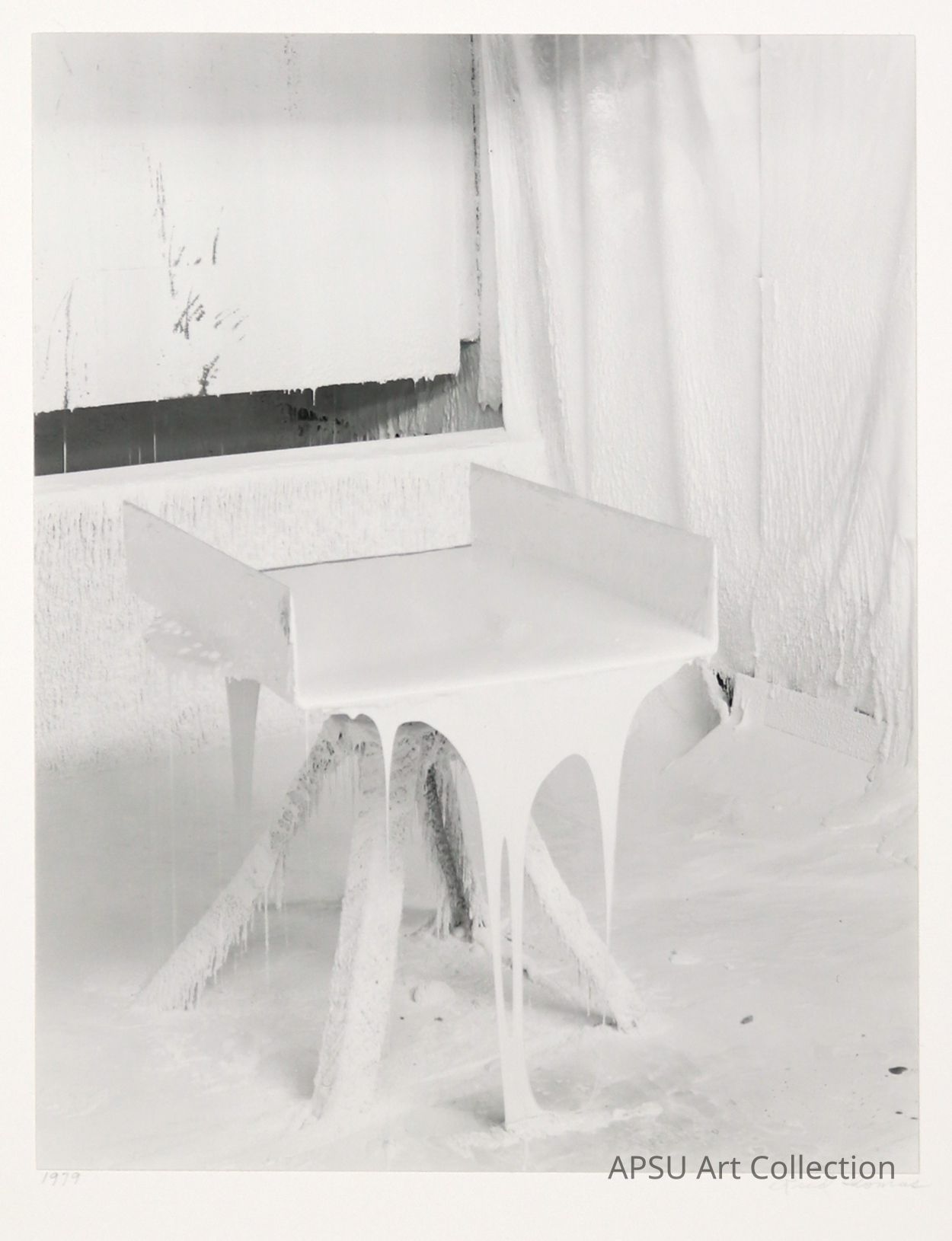 This image portrays a minimalist scene featuring a modern, inverted U-shaped desk covered with what appears to be thick, textured layers, possibly paint or a plaster-like substance, contributing to a surreal, frosty aesthetic. The desk is positioned against a backdrop exhibiting similar textures, and the setting's monochromatic tone enhances a sense of abstract coldness, almost as if the scene is frozen in time.