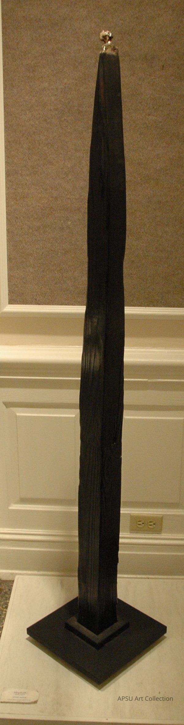 Multipet: The image shows a tall, slender sculpture with a dark, textured surface standing on a square base in an indoor setting. The sculpture narrows at the top and features a small metallic element, giving it a modern, abstract appearance. Its base is simple and geometric, enhancing its verticality.