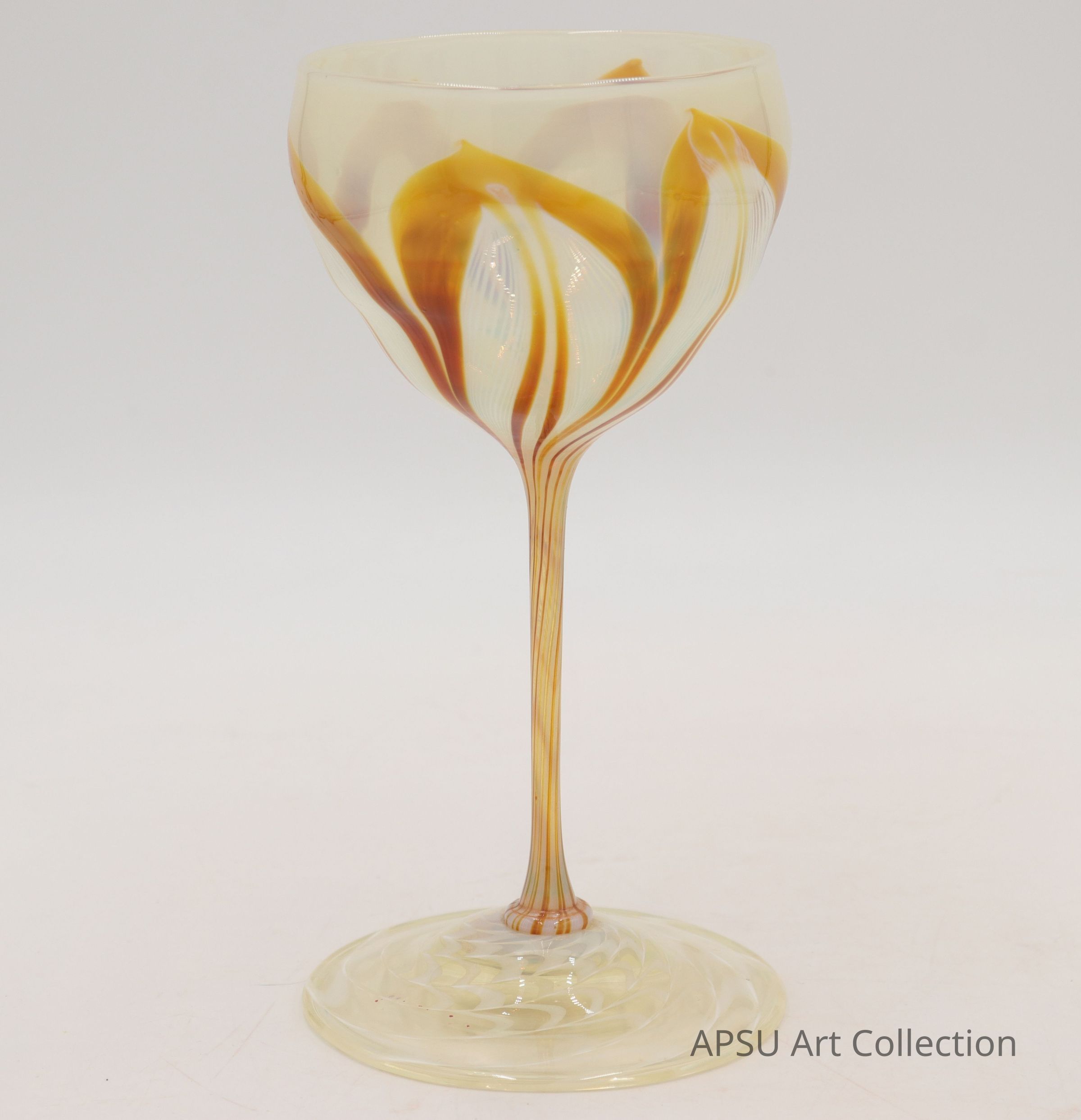The image shows an elegantly designed goblet, crafted with intricate glasswork patterns in shades of amber and cream. The stem of the goblet is slender and delicate, featuring swirling lines that extend from the base to the bowl. The base itself is beautifully rounded, providing a sturdy foundation for the piece. These warm tones create an appealing aesthetic, accentuating the graceful curves of the glass. The patterns on the bowl mimic the natural flow of liquid, enhancing the overall visual appeal. This piece combines artistry and