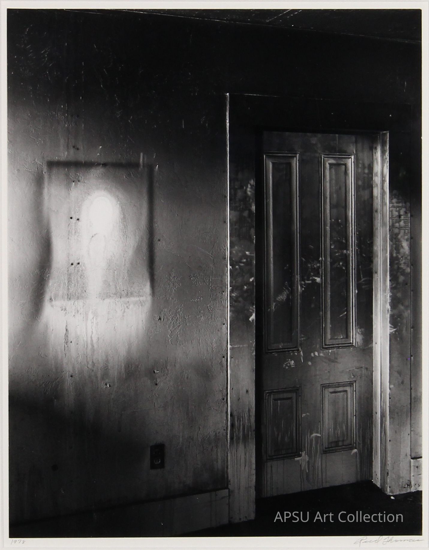 The black and white photograph presents a texturally rich, shadowy room featuring an open double door on the right side, through which some exterior light spills in, contrasting with the darker interior. On the left, a mysterious, eerie silhouette of an apparent figure, resembling a ghostly presence, is captured within a lighter frame made by a doorway, adding a surreal and haunting quality to the scene. The walls and the floor are worn and marked, enhancing the overall mood of decay and neglect.