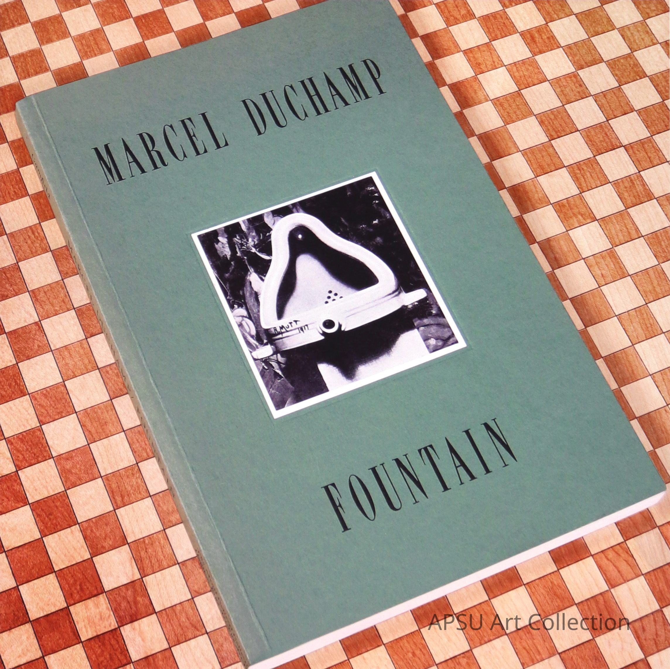 The image features a book with a seafoam green cover titled "Marcel Duchamp" at the top and "Fountain" at the bottom. In the center, there is a black and white photograph of Duchamp's famous art piece, a porcelain urinal, displayed against a contrasting patterned background of a brown and cream checked background.