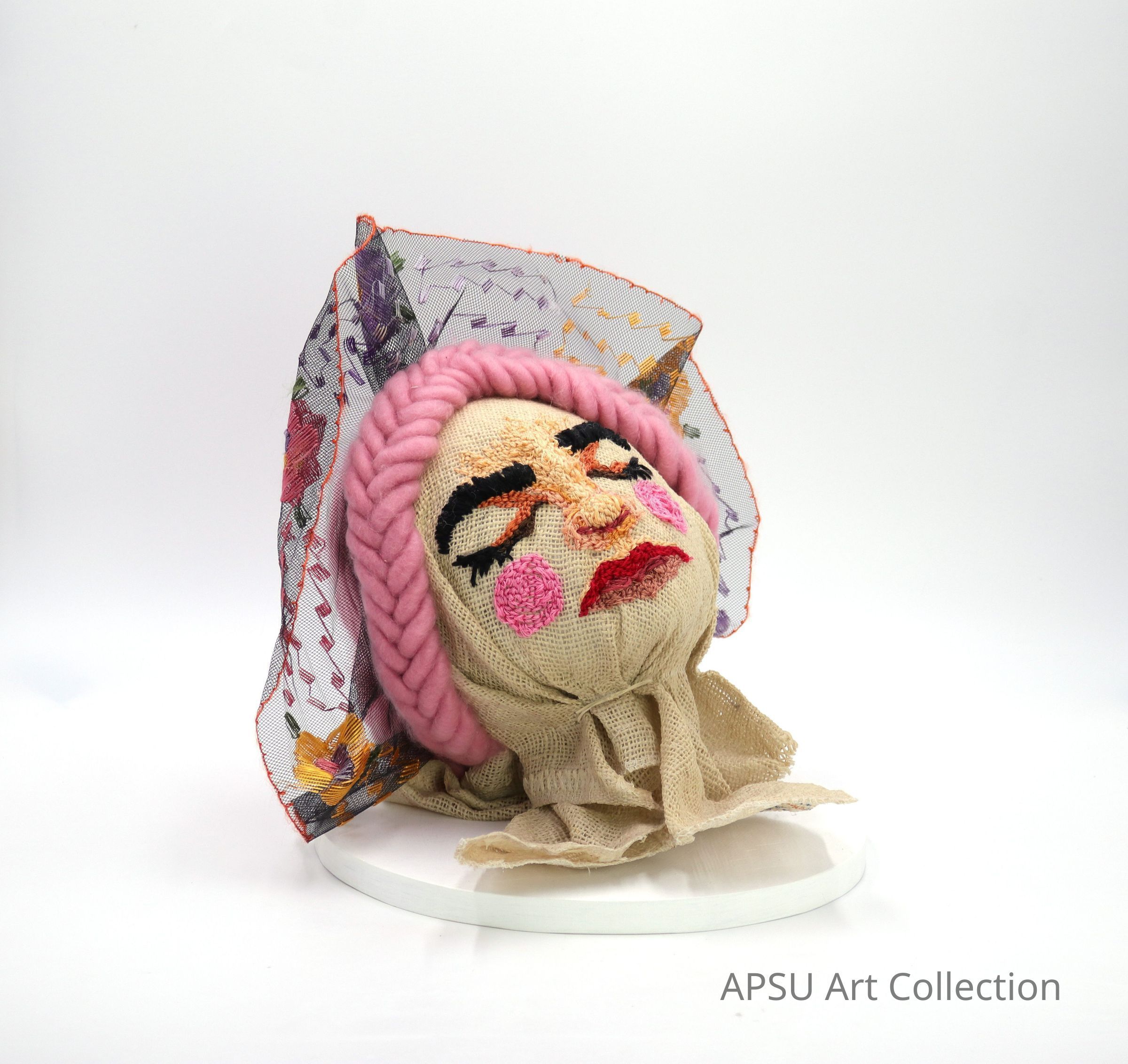 This image showcases a visually intriguing sculptural piece, featuring a textile-based head adorned with pink yarn hair and a textured, colorful scarf elegantly draped around it. The face is detailed with embroidered features including closed eyes with long lashes, pink cheeks, and vibrant red lips, presenting a serene and crafted artistic expression. The piece is mounted on a white stand against a plain background, highlighting its vibrant colors and intricate textures.