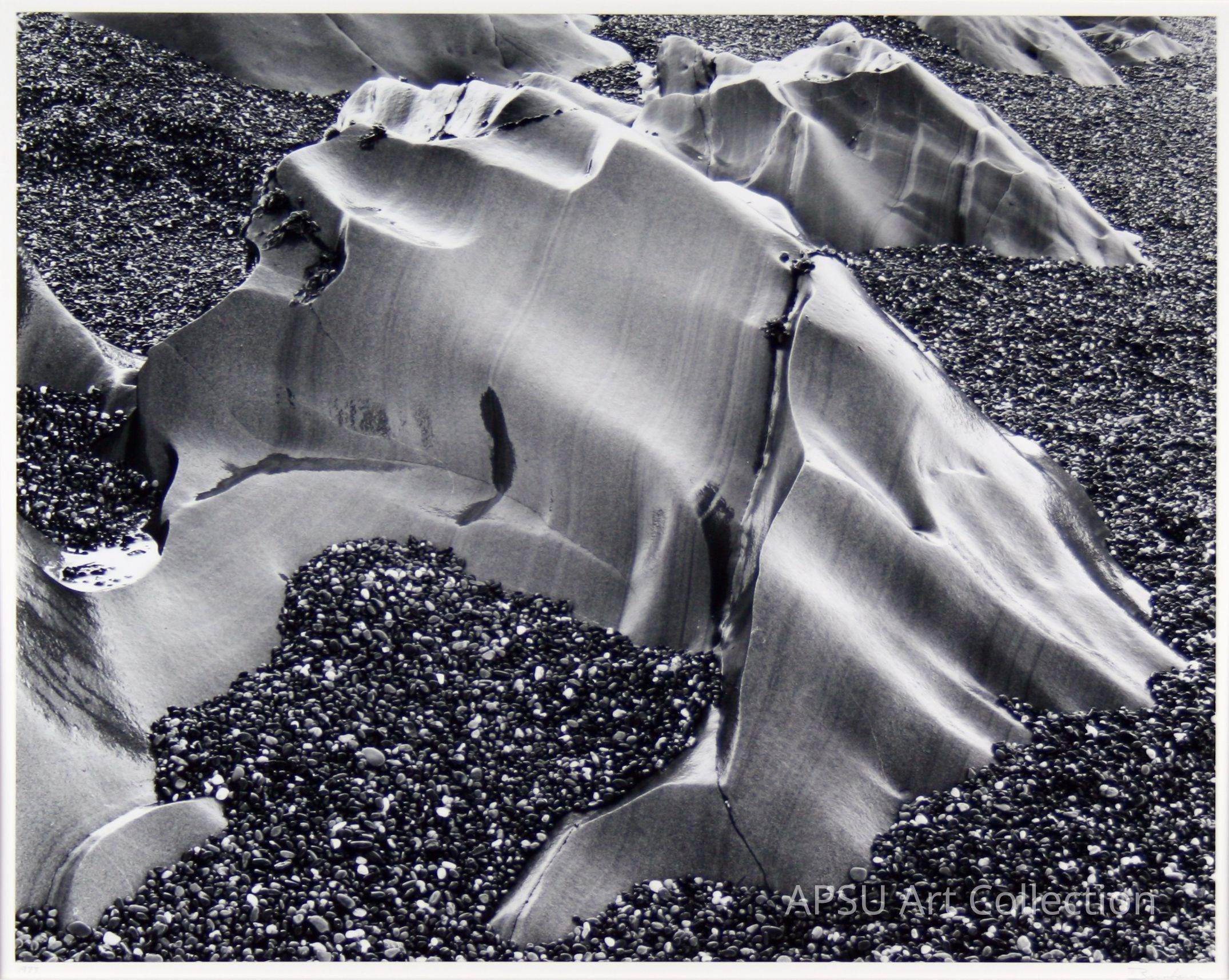 This black and white photograph captures a striking landscape of smooth, wind-carved sand dunes, contrasting sharply with the surrounding areas covered in small, dark pebbles; the intricate textures and shadows emphasize the natural beauty and erosion patterns of the dunes, creating an almost otherworldly atmosphere that invites contemplation of the forces that shaped this scene over time.