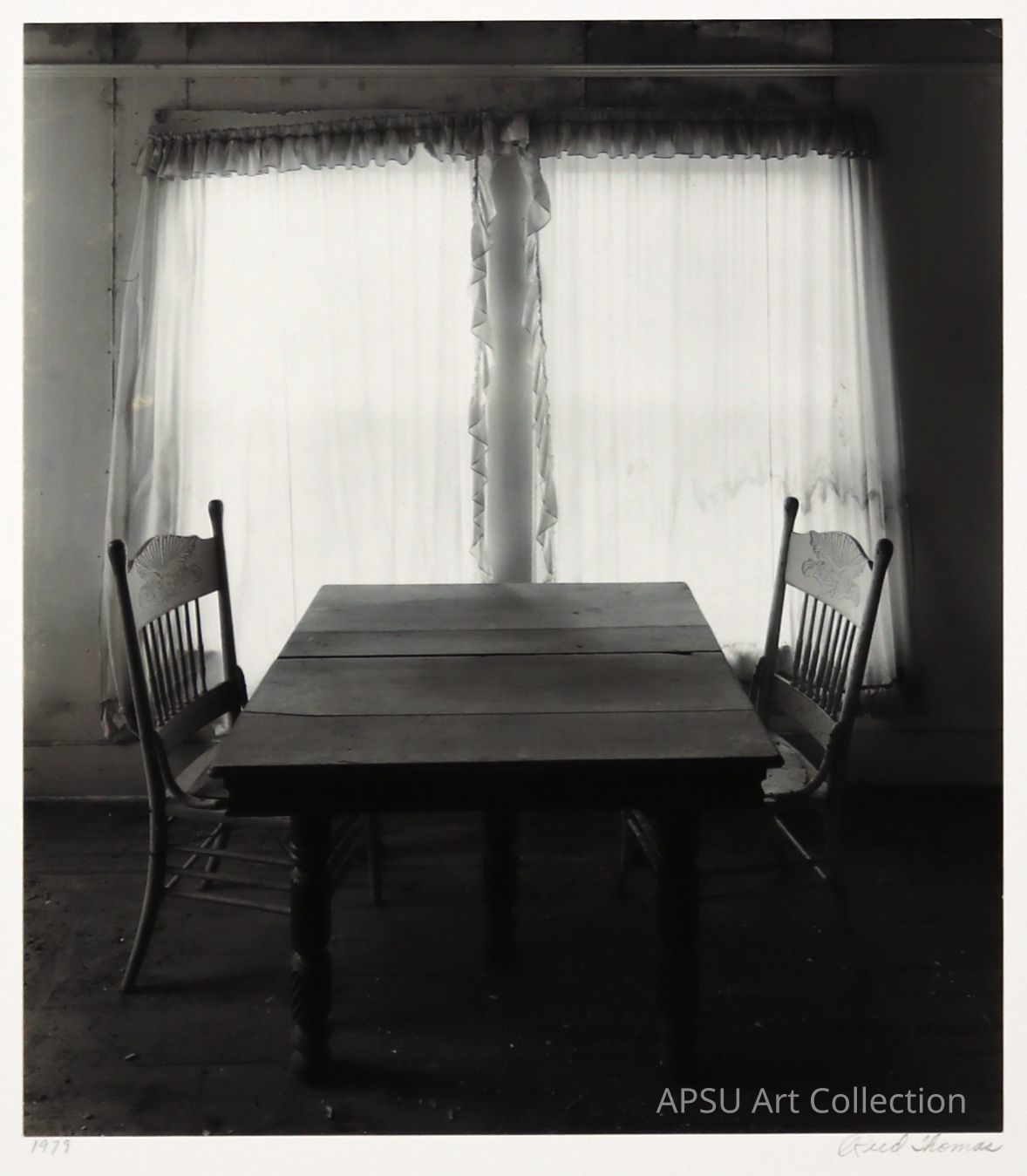 The black and white photograph captures a sparsely furnished room featuring an aged wooden table flanked by two traditional chairs with ornamental designs carved into their backrests. Positioned against a curtained window that allows muted light to seep through, the setting conveys a stark, serene simplicity typical of rustic interiors, evoking a sense of quietude and the lingering presence of past conversations.