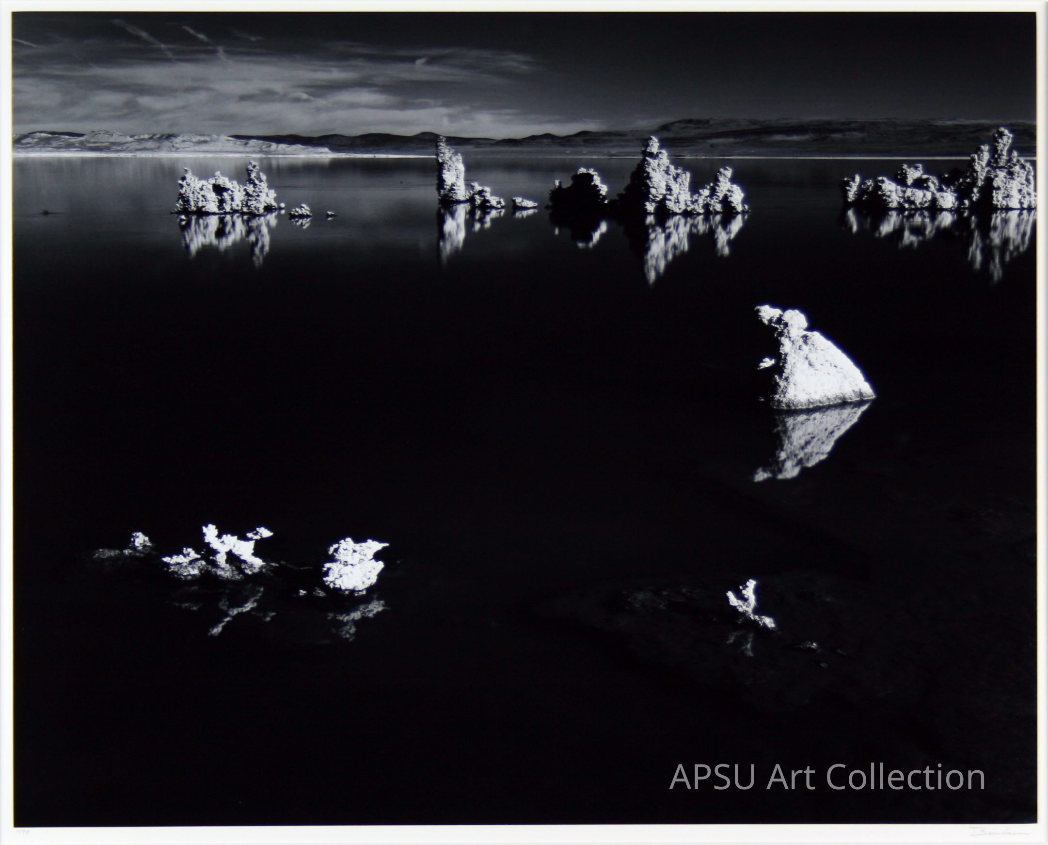 The black-and-white photograph features a serene and reflective body of water surrounded by stark, rugged formations that jut out of the lake's surface, their rough textures contrasting sharply with the stillness of the water, which mirrors the formations and the distant horizon. The sky, stretched out above, hints at motion with its streaking clouds, adding a dynamic element to an otherwise calm scene, while the distant mountains provide a timeless, immutable backdrop that anchors the composition.