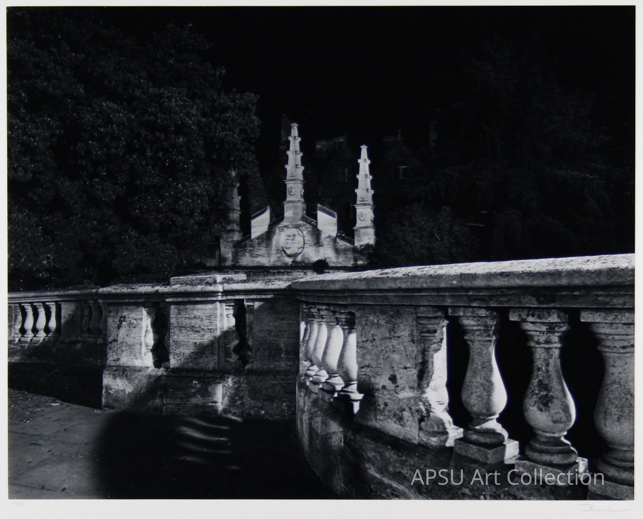 In the black-and-white image, an architectural structure with intricate stone balustrades is prominently featured in the foreground, showcasing detailed craftsmanship through its textured columns and ornate patterns, evocative of historical or classical design influences. In the background, two tall, slender towers stand on either side of an elaborate central facade, which is adorned with a circular emblem that adds an element of grandeur and symbolism to the scene.