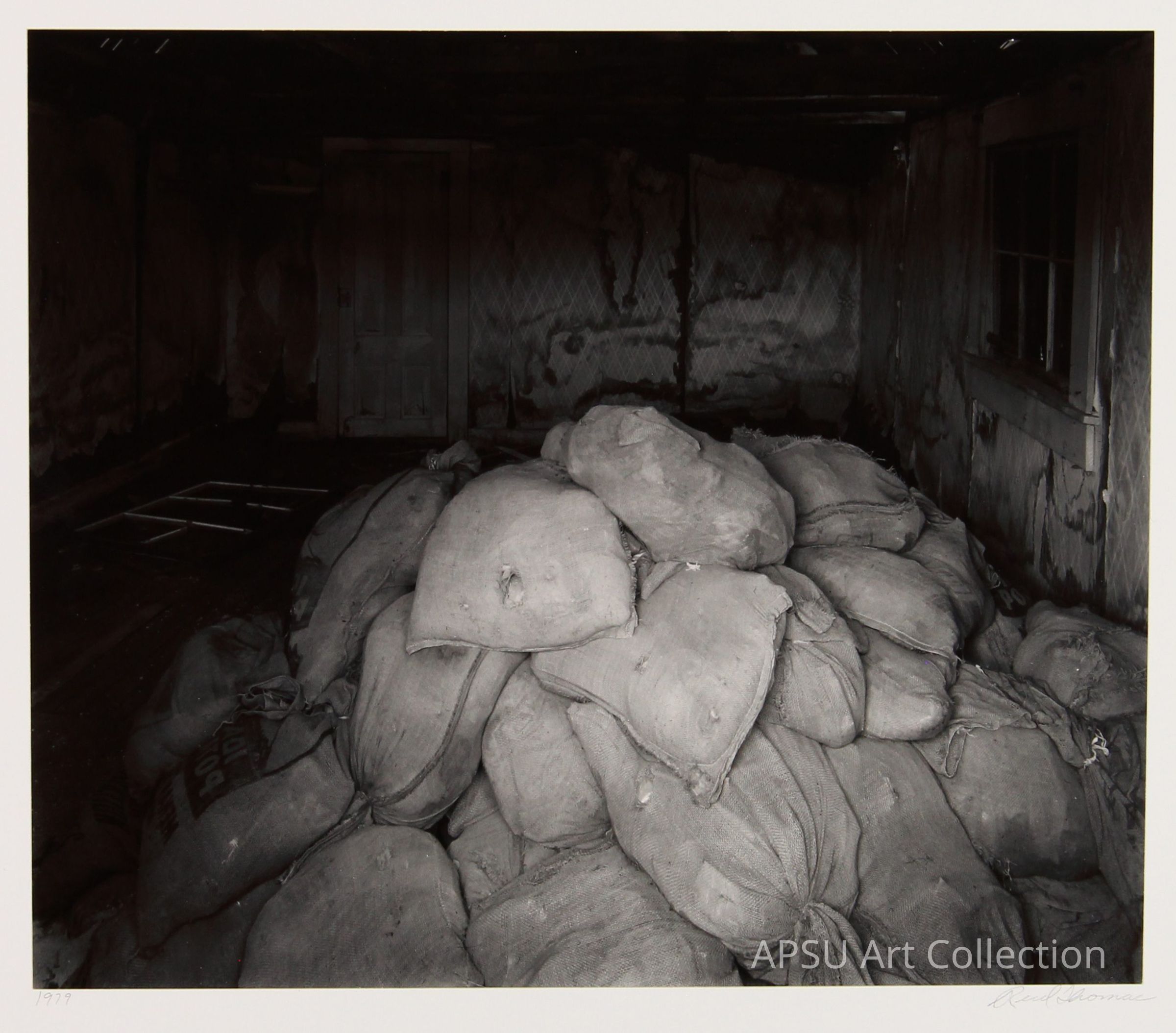 This black-and-white photograph captures a dimly lit, deteriorating interior, possibly a storage space, cluttered with a large pile of what appear to be weathered and worn burlap sacks. The room features peeling walls, a decrepit wooden door, a grimy window allowing minimal light, and stark shadows cast across the scene, depicting desolation and neglect. The composition, lighting, and subject matter evoke a sense of abandonment and perhaps historical significance.