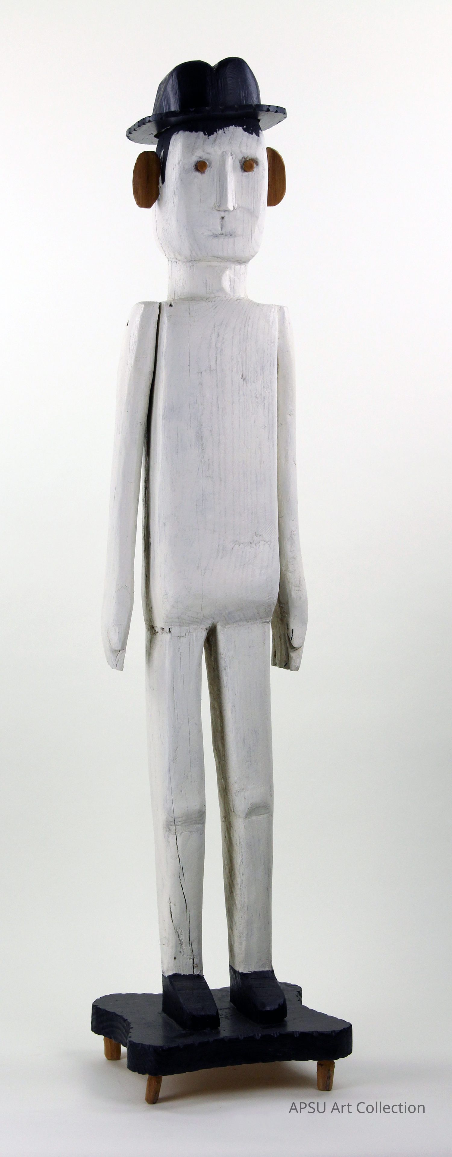 The image depicts a tall, slender wooden sculpture of a human-like figure painted in a muted gray-white palette, standing upright on a black platform supported by pegs. The figure features oval-shaped, peanut-colored ear details and orange eyes, dressed in a simple long-sleeve top and pants, complemented by a black hat, creating a minimalist yet expressive artistic representation.