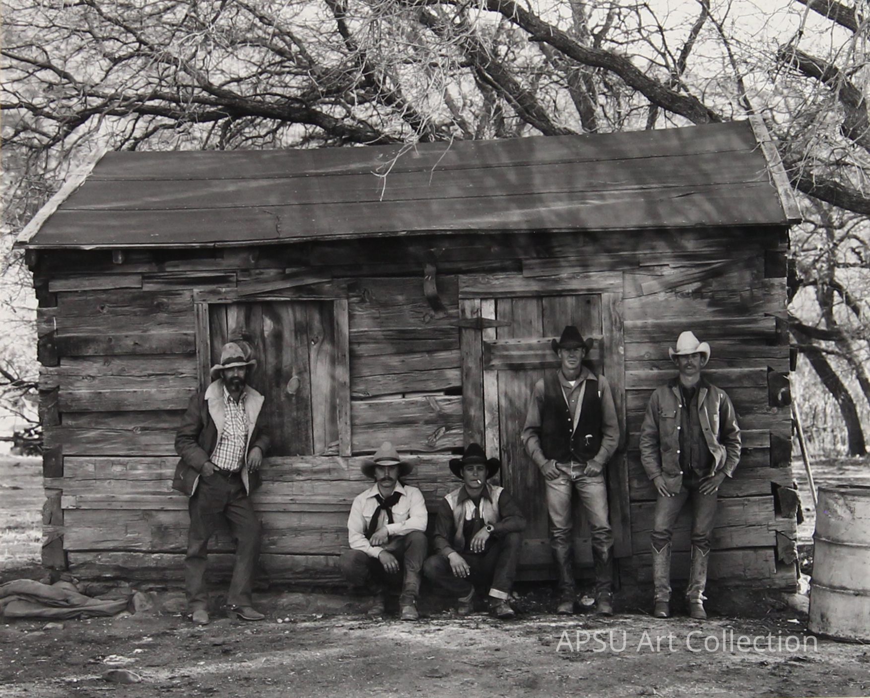 The black-and-white photograph captures five men dressed in traditional Western attire, each exuding a distinct rugged charm. They stand and sit in front of an aged, wooden cabin, which sits under leafless trees, suggesting a serene, rural setting. Their attire includes cowboy hats and boots, and their relaxed poses combined with their serious expressions evoke a sense of camaraderie and quiet strength, typical of classic Americana.