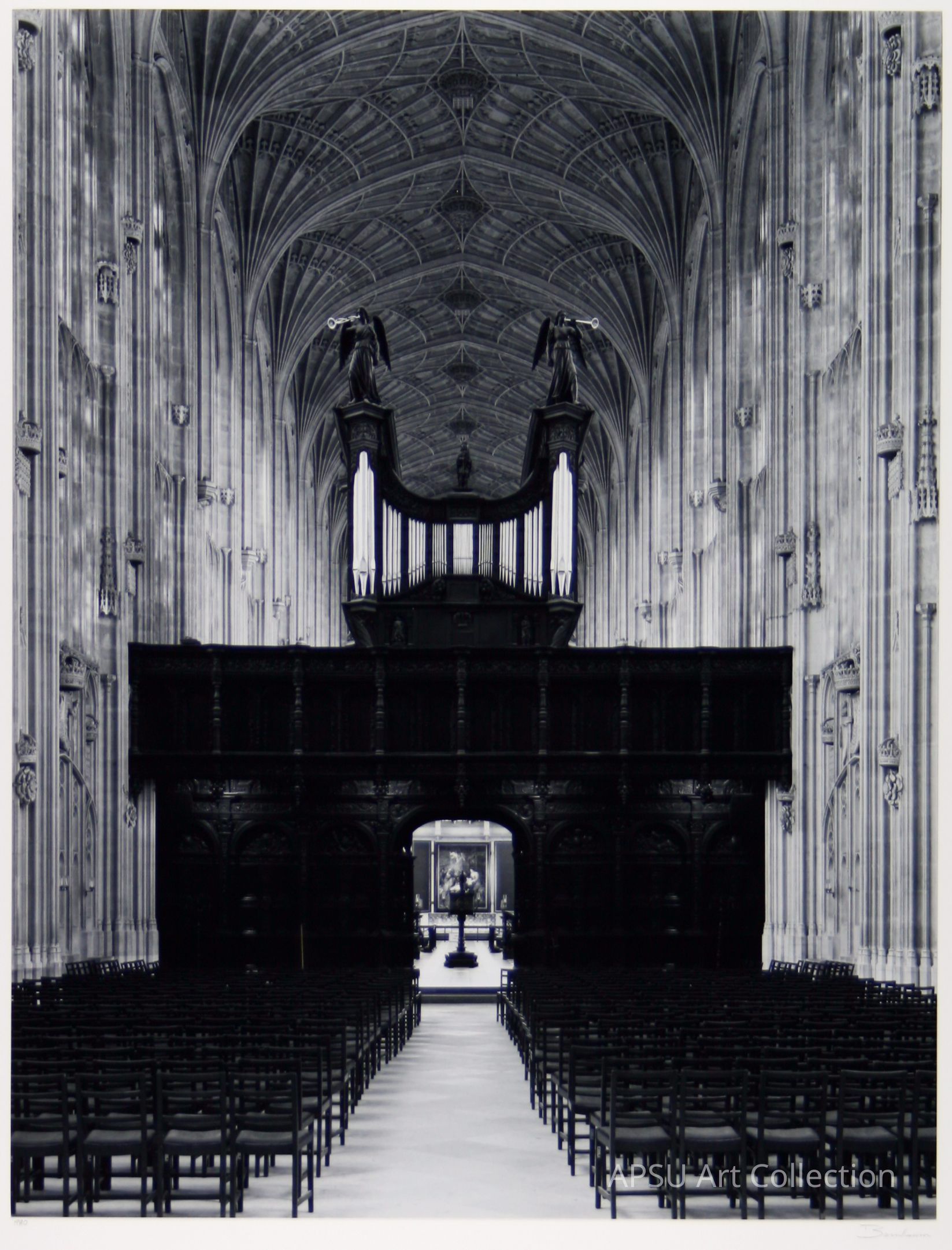 The image depicts the interior of a grand, Gothic-style cathedral with elaborate fan vaulting on the ceiling, intricately carved stonework adorning the tall walls, and an expansive aisle flanked by rows of empty chairs. In the center, an imposing organ with tall pipes stands above a dark, ornately designed wooden screen that stretches across the width of the space. The symmetrical design draws the eye towards the altar area at the far end, partially visible through the archway, highlighting a beautifully lit artwork.