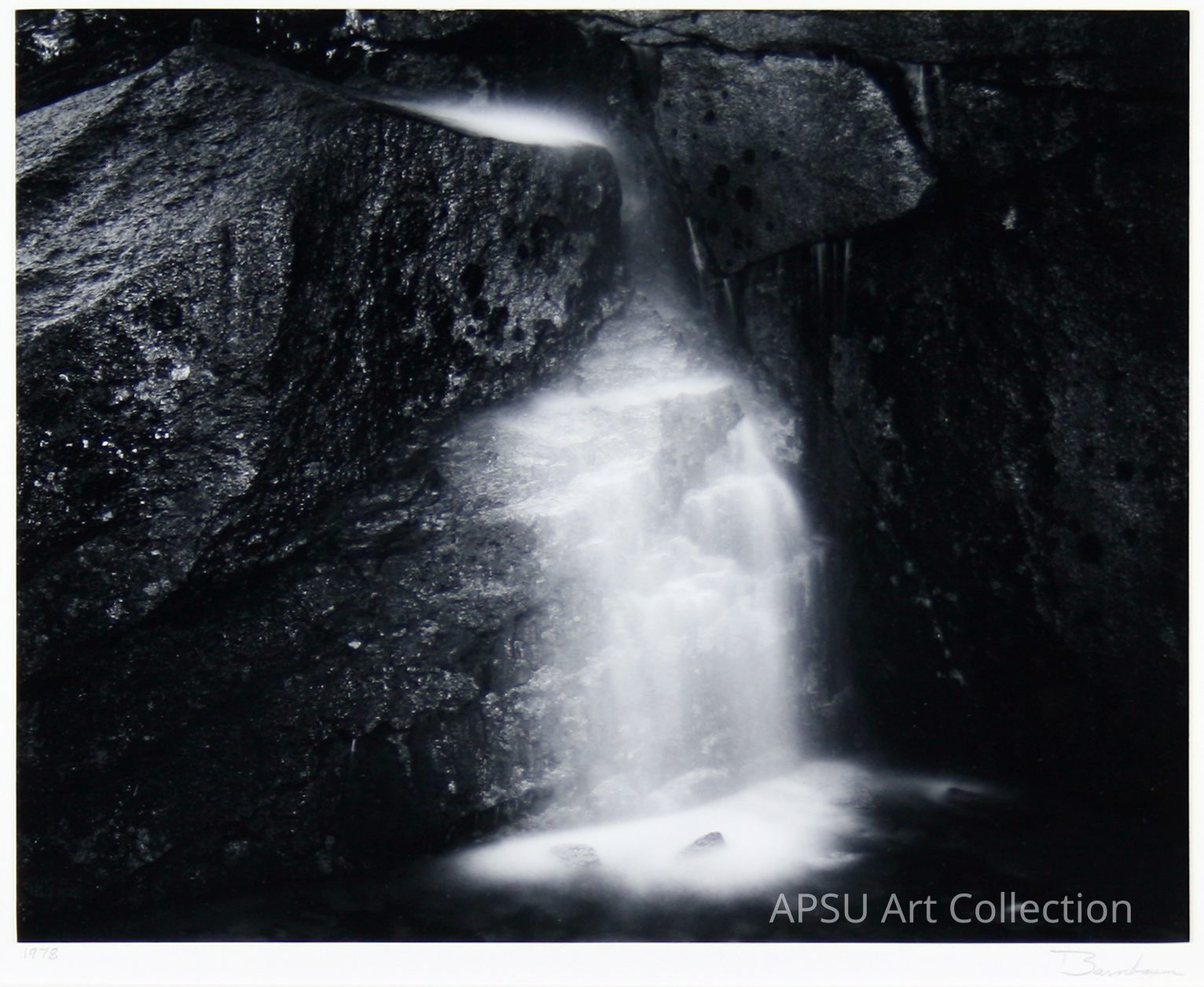 The image exhibits a captivating black and white photograph of a serene waterfall cascading over rugged, textured rocks, creating an ethereal, misty effect as the water flows smoothly downward; the stark contrast between the dark, almost ominous rocks and the soft, bright water highlights the natural beauty and tranquil essence of this secluded scene, capturing the moment where water meets stone in a harmonious yet dynamic balance, suggesting almost a timeless quality to the interaction of these elemental forces.