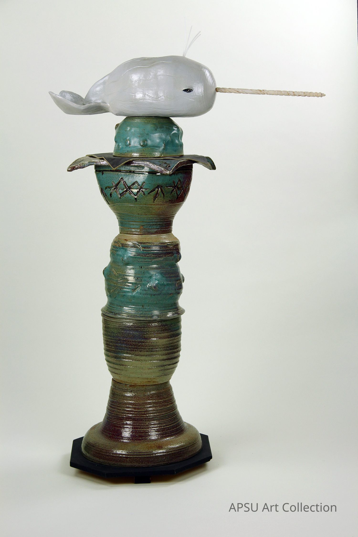The image displays a unique sculpture featuring a stylized narwhal with a long, pointed horn, positioned atop a tall, intricately designed pedestal. The pedestal is composed of various textures and colors, including shades of brown and green, with carved patterns and ridges that create an artistic and visually engaging structure. The narwhal, painted in a light gray color, contrasts with the earthy tones of the pedestal, drawing attention to its smooth, rounded form and the delicate detailing of its horn.