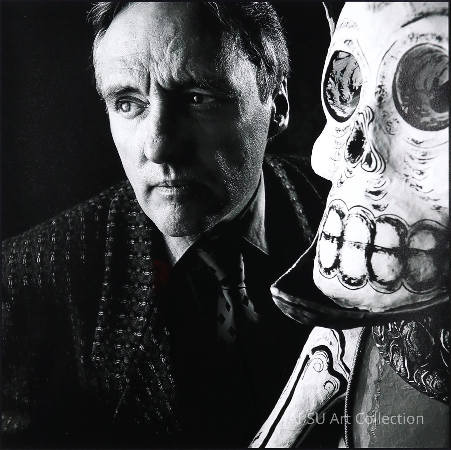 The image depicts a middle-aged man, looking intently towards his left, standing next to a large, ornate skull, creating a striking contrast and suggesting themes of mortality and introspection. The man's expression is somber and reflective, enhanced by the monochrome palette, which adds a dramatic and timeless quality to the scene. His attire includes a textured jacket and a patterned tie, suggesting a formal occasion or personal significance to the moment captured. The skull itself is detailed, with visible sutures