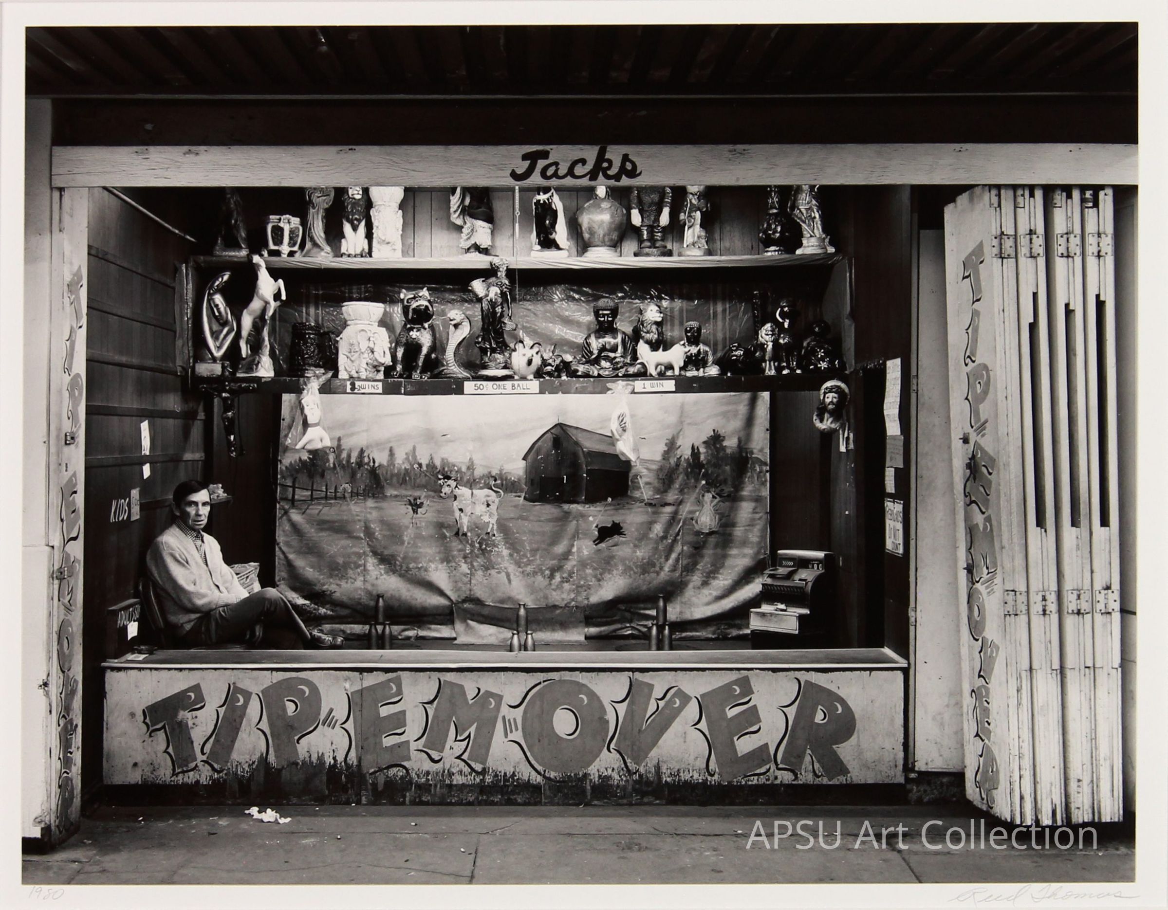 The black and white photograph shows a man sitting pensively in a kiosk named "Jacks" decorated with various items and dolls, under a sign that reads "TIP EM' OVER". The booth features prizes displayed on shelves and a rural landscape painting, capturing a slice of Americana, likely at a fairground. This scene, possibly from the mid-20th century, emerges with a candid and atmospheric quality, reflecting a particular moment in everyday life.