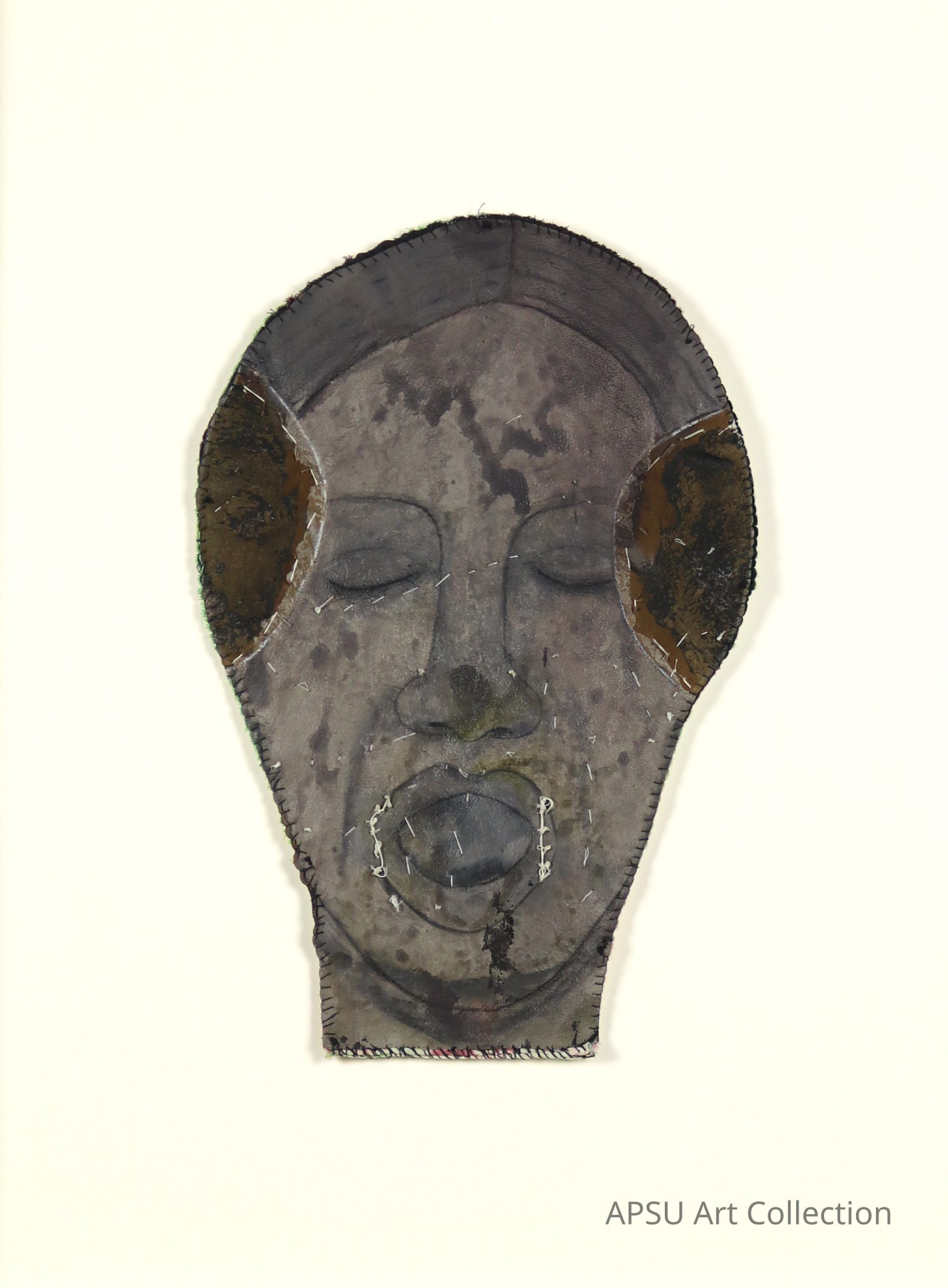This image features a textured, portrait-style representation of a face, appearing as a crafted mask with subtle, monochromatic tones and noticeable stitched details. The face is serene, with closed eyes and lips sewn shut, framed by what appears to be dark, rough patches that suggest hair or organic textures. There is also notable damage or deterioration at the bottom of the mask, adding a sense of age or decay to the artwork.