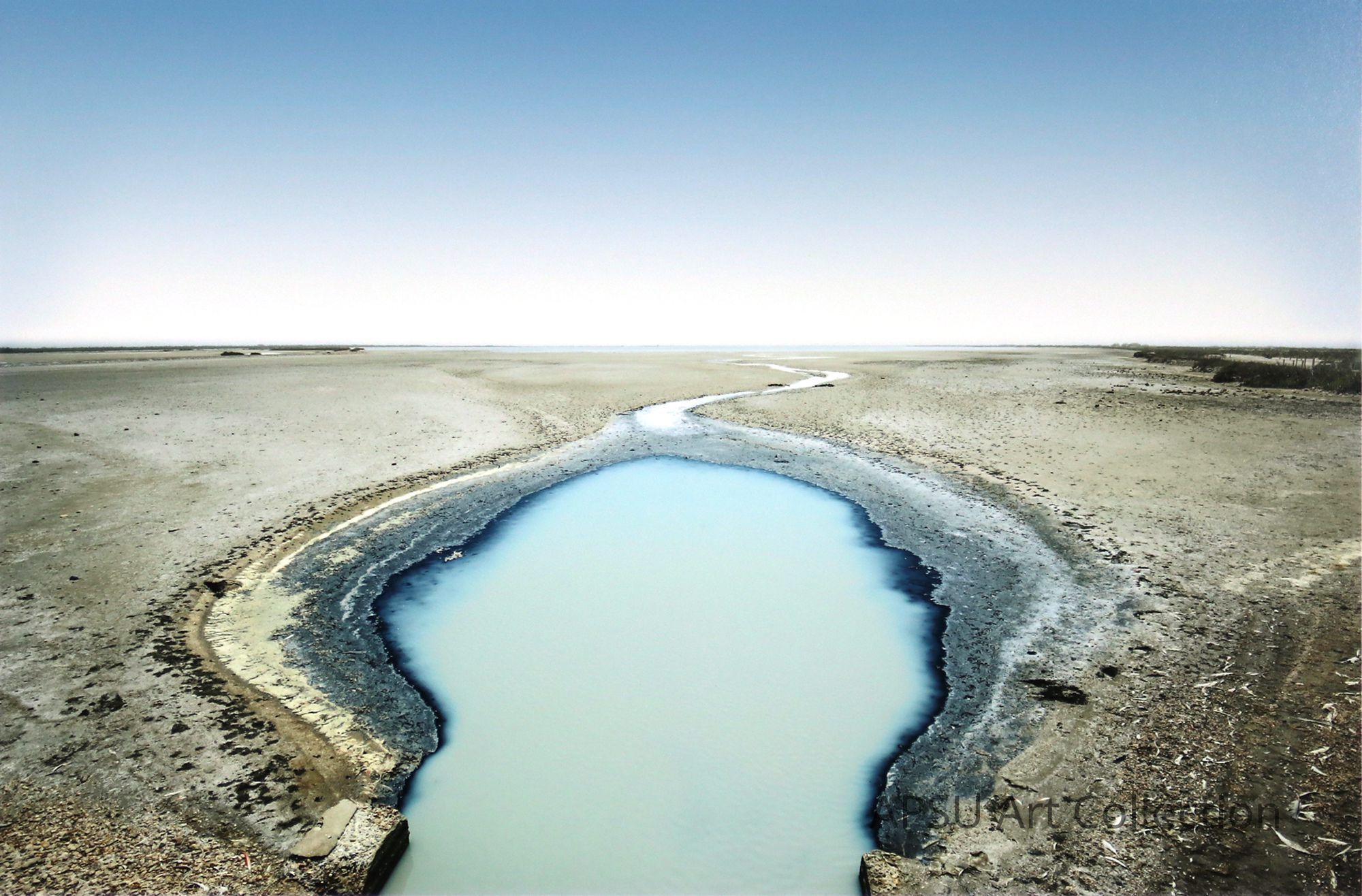 The image captures a serene, expansive landscape, predominantly featuring a vast, flat terrain with a distinctive blue water channel meandering through it. This waterway, resembling a slender, winding river, contrasts starkly with the surrounding muted earth tones of the dry, cracked soil and sparse vegetation. The clear sky merges seamlessly at the horizon, adding to the desolate yet tranquil ambiance of this seemingly remote area, possibly a tidal flat or salt pan in a coastal region.