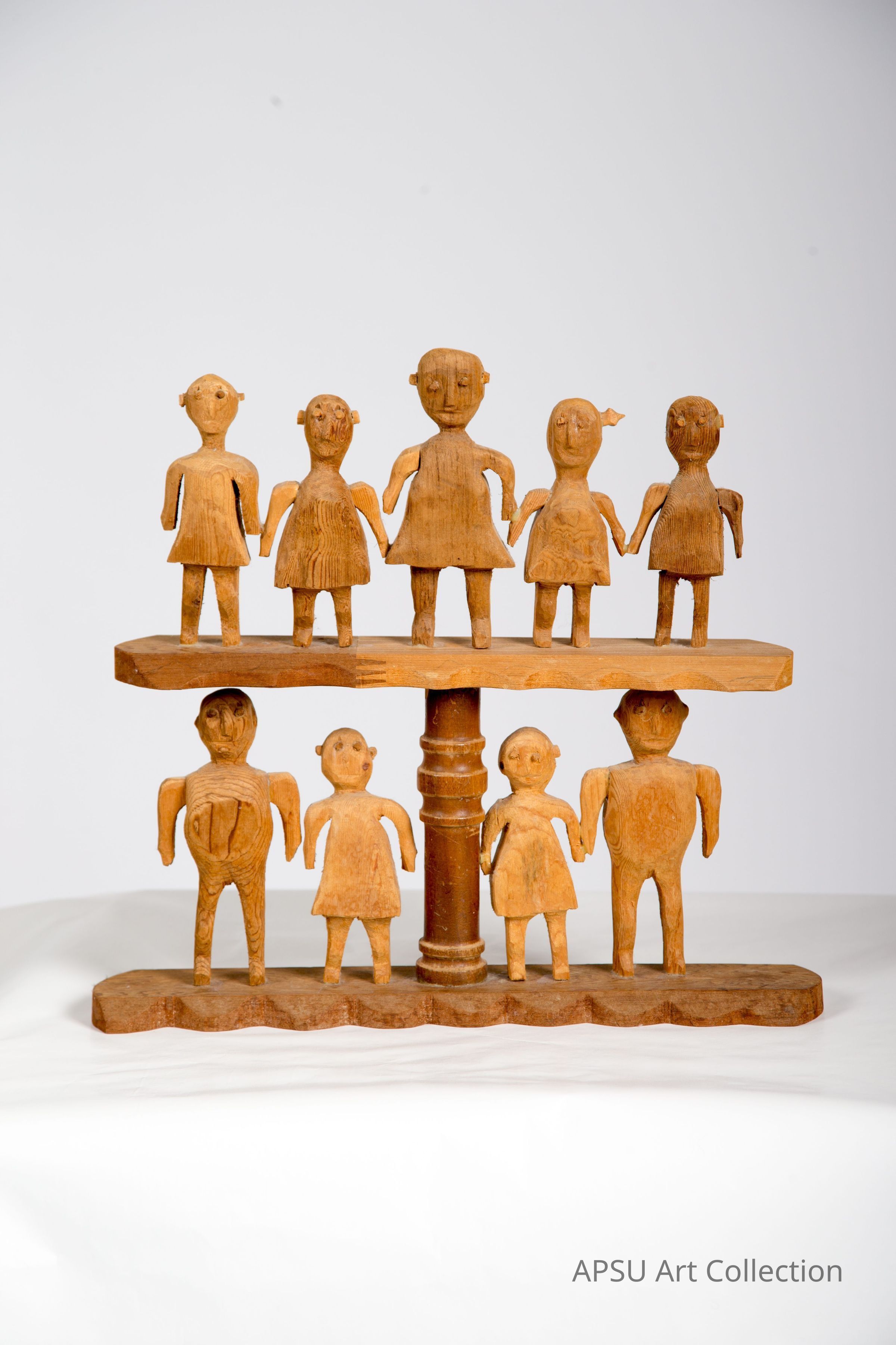 The image shows a tiered wooden sculpture, consisting of a central, vertical spindle supporting two circular platforms, each displaying a group of hand-carved, stylized human figures. The upper platform hosts five figures and the lower platform holds four, all crafted with a rustic finish and distinct, simplistic features that suggest an artistic or folkloric theme, likely making this piece an example of traditional craftsmanship or contemporary art. The plain white background enhances the focus on the wooden texture and the forms of the figures.