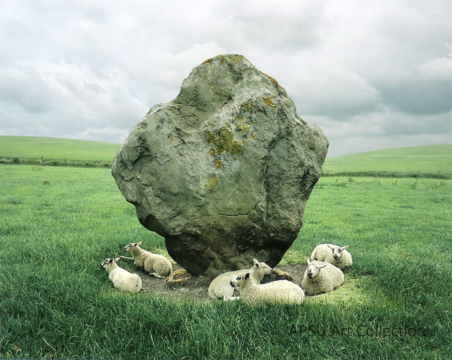The image shows a group of lambs resting in the shade of a large, moss-covered boulder in a lush green pasture under a cloudy sky. The vast, open landscape extends to the horizon, conveying serenity and the simplicity of pastoral life. The setting seems calm with the lambs comfortably huddled together, suggesting a quiet, peaceful day in a rural environment.