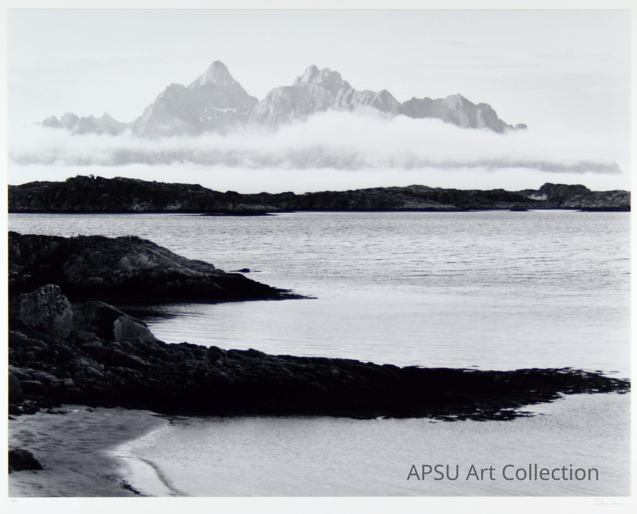The image depicts a serene black-and-white landscape featuring a calm body of water in the foreground with rugged, rocky shores, while in the background, towering mountains emerge majestically, shrouded partially by a layer of mist or low-lying clouds, creating a dramatic and moody atmosphere that evokes a sense of tranquility and solitude.