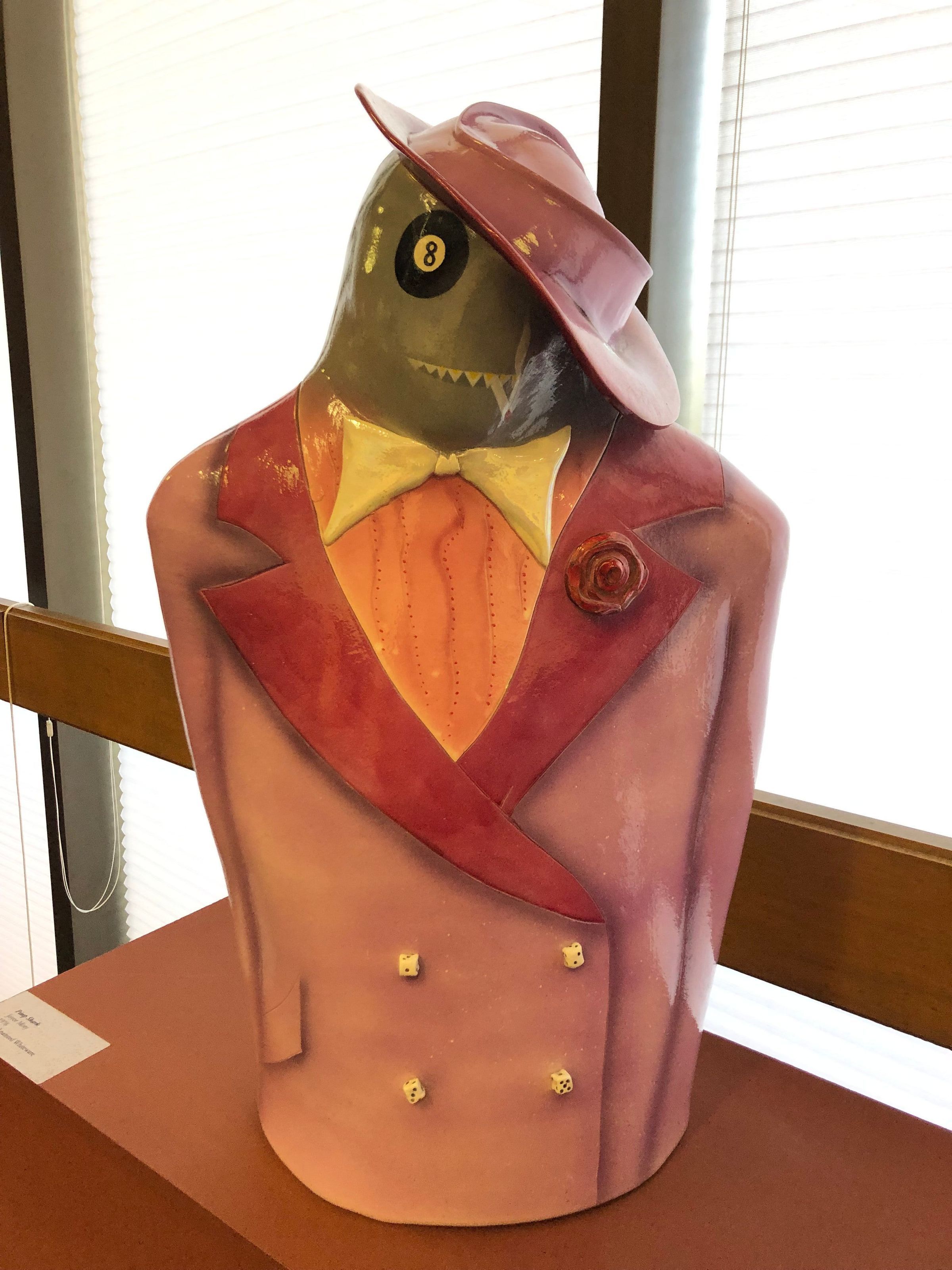 Shark Pimp: Medium sized ceramic sculpture of a grey shark with a tuxedo and hat. The tuxedo is a lavender base with a mauve pink collar. There are 4 dice painted buttons on the lower torso section. The back side is covered with black dominoes with white dots leading out to the right shoulder. Grey and white music notes are also located on the right shoulder with two martini cups below it, a green olive located in each.