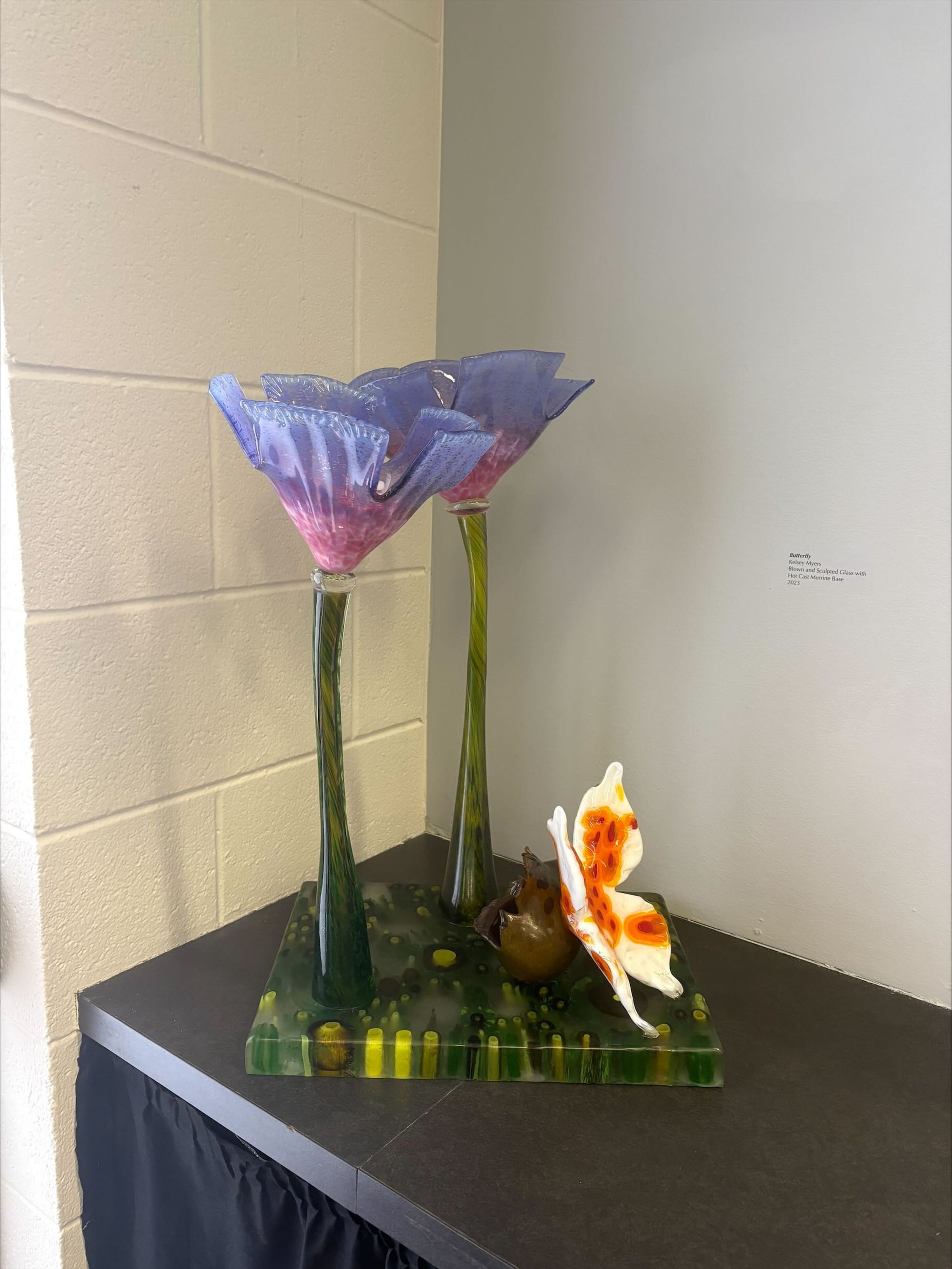 Butterfly: A large free-standing glass sculpture that depicts imagery of flora and fauna. The work has a Muti-colored green and yellow base with two Muti-colored pink and purple flowers and a Muti-colored white and orange butterfly that sits next to the flowers.
