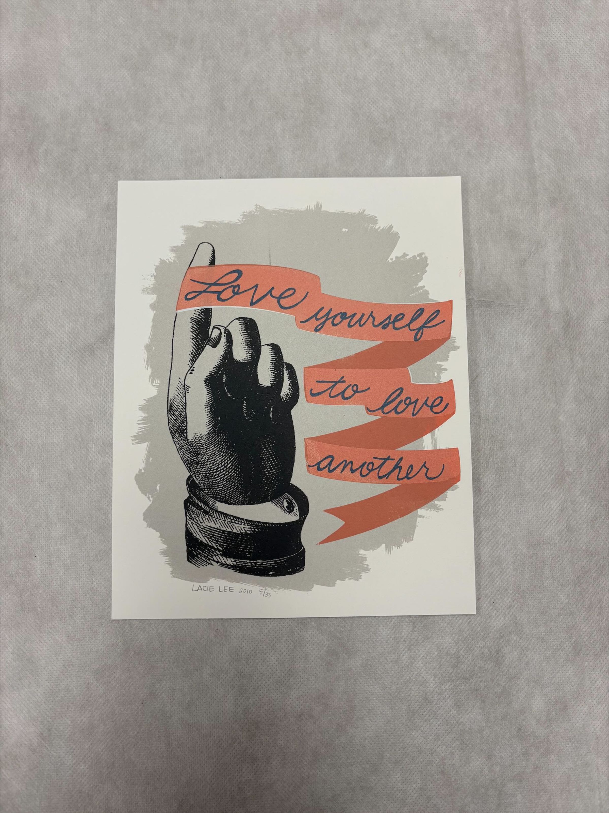 Front: A vertically oriented, medium sized print that uses black, grey , peach and blue pigment. The print contains a central image of a hand that has a ribbon with text tied to its finger that loops down towards the bottom of the print.