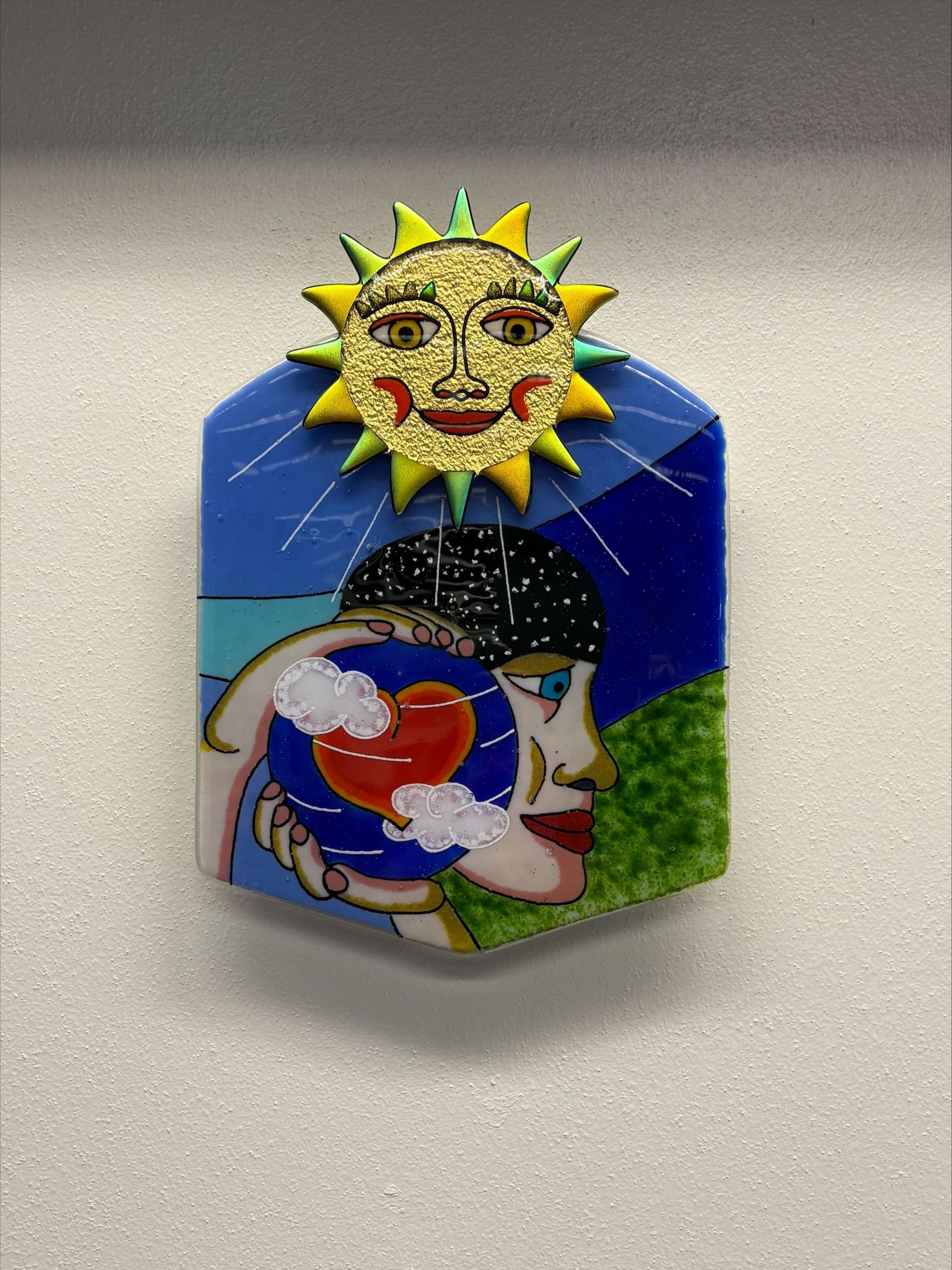 Condition Photo: A medium-sized, rounded, hexagonal plate of fused glass with multicolored design. This plate features a sun at the top center of the plate with figural imagery of a person holding a blue sphere details with a heart and clouds taking up the middle section of the plate.