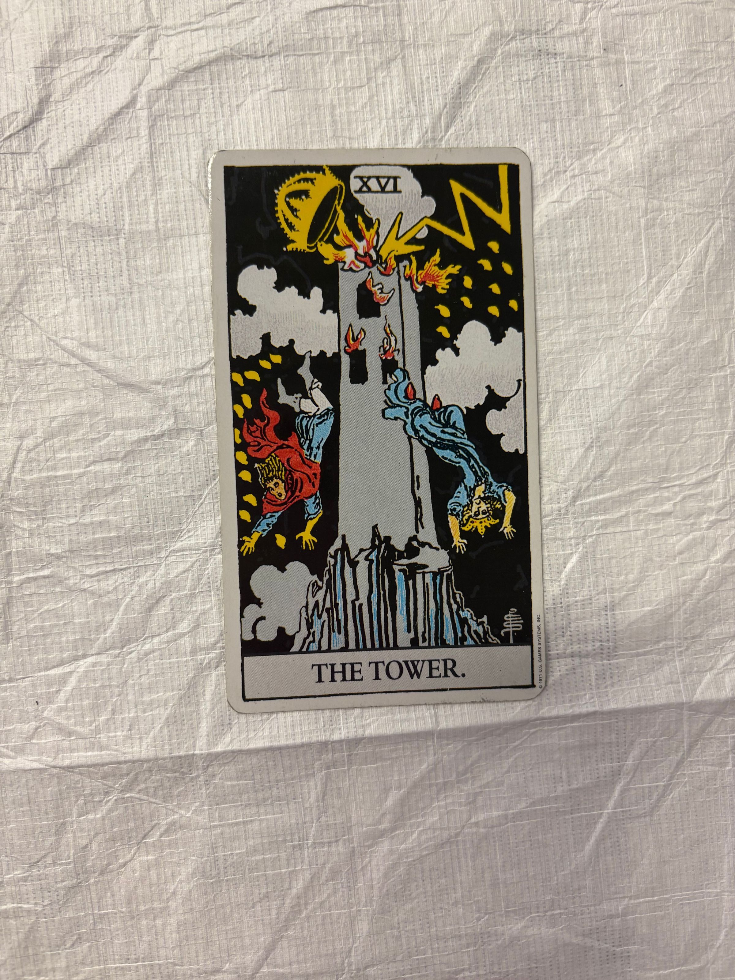 Front: A small, vertically oriented print that mimics the look and design of a taro card. The print utilizes both imagery and text along with multiple colors. At the center of the print is a large tower that is on fire and from that burning tower jump two figures that can be seen falling on both sides of the tower.