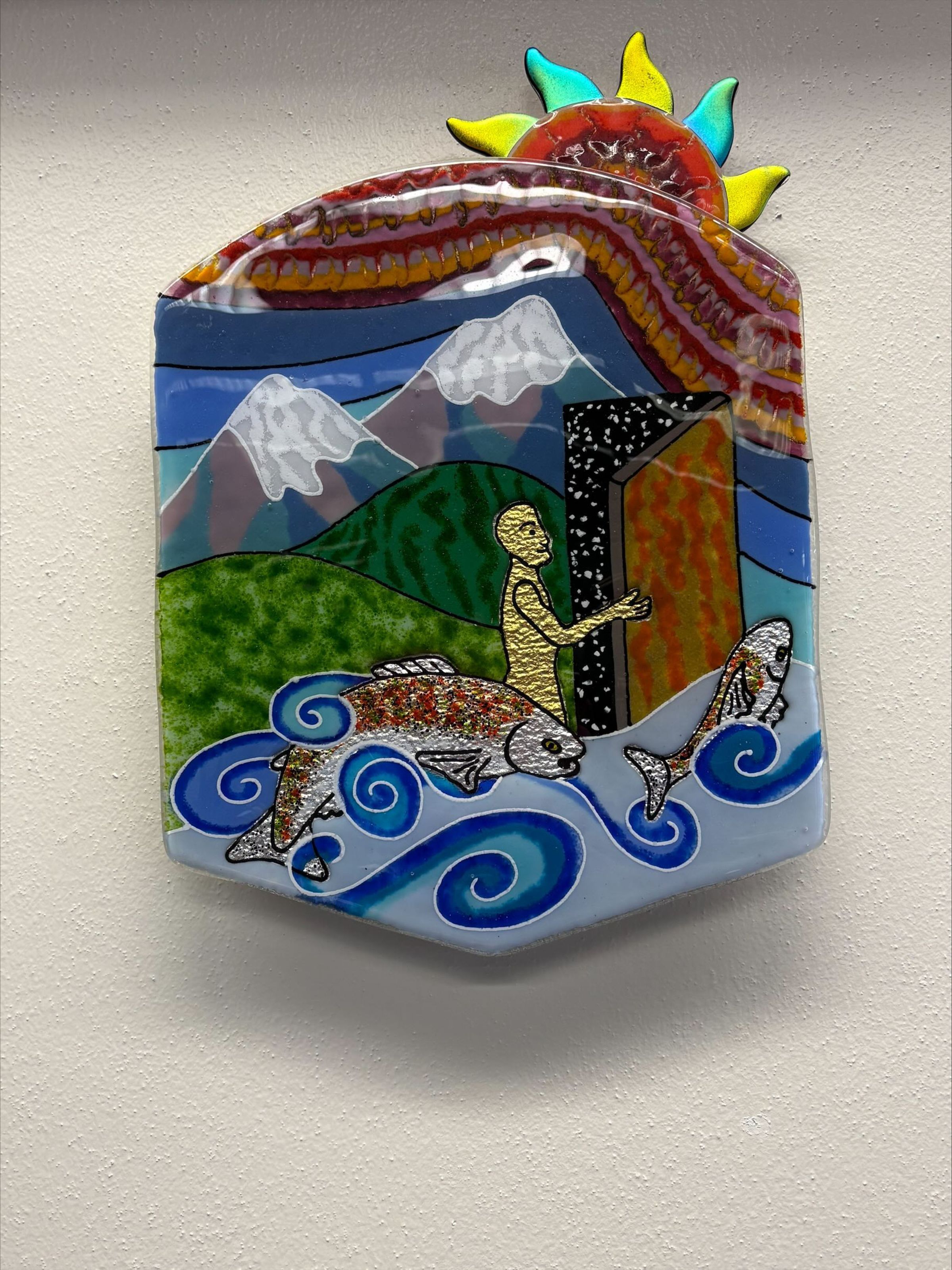 Condition Photo: A medium-sized, rounded, hexagonal plate of fused glass with multicolored design. The plate has an image of a sun attached to the top proper-left corner, and also has images of a distant mountain landscape, a figural form opening a strange door and a river with fish.