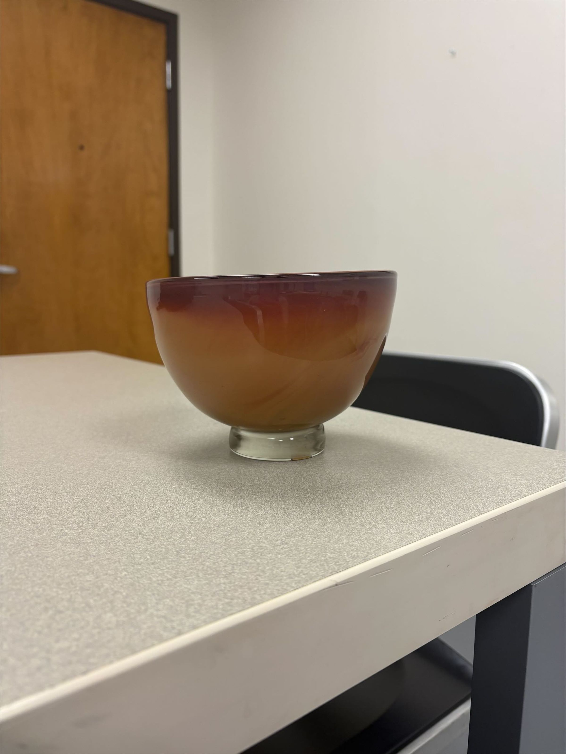 Condition Photo: A small multi-colored handblown glass bowl that has a subtle red/brown color gradient. The bowl is not uniformly shaped and tilts upwards on one side.