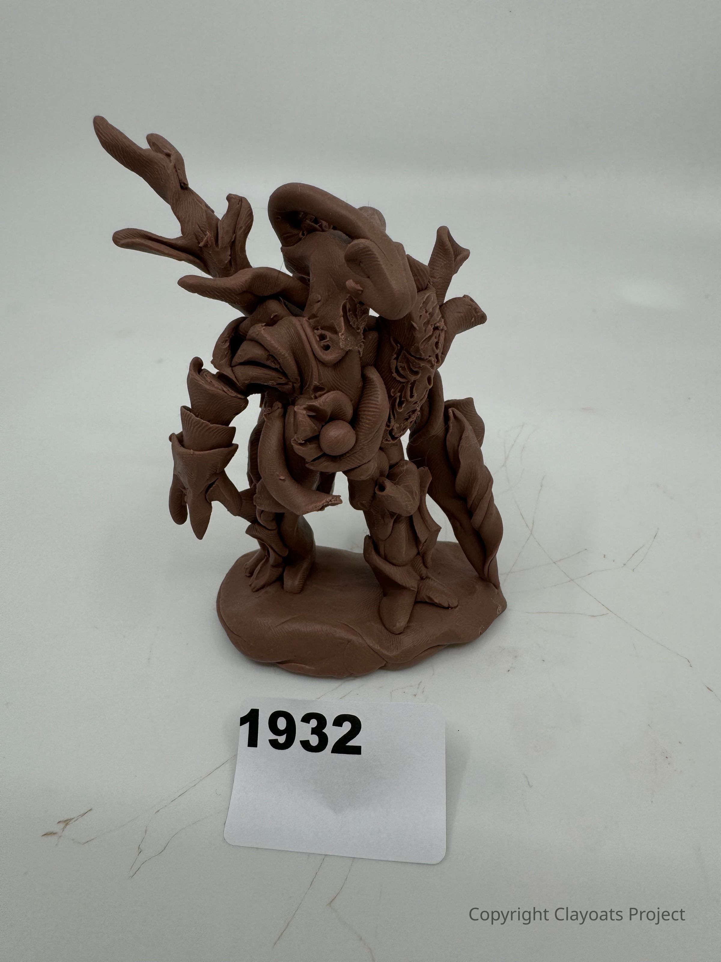 The image depicts a clay sculpture with intricate, abstract forms and textures, featuring several twisting and branching appendages that give it a dynamic and organic appearance. The sculpture stands on a flat base, The overall color of the sculpture is a natural, earthy brown, and the background is a neutral off-white, allowing the details of the artwork to stand out prominently.