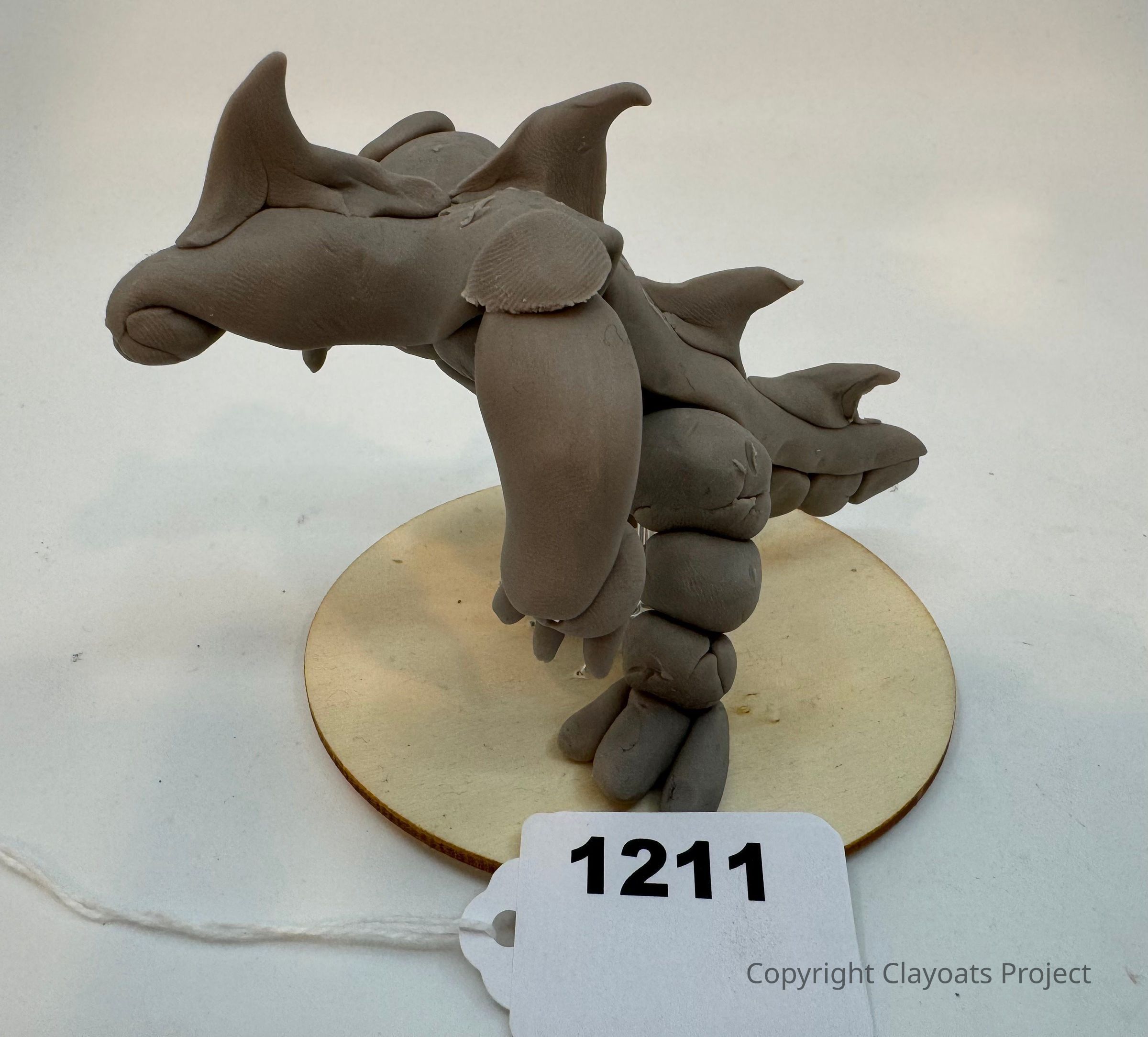 The image shows a gray sculpted creature made from clay, featuring multiple horns and a set of legs, positioned on a circular wooden base ; the surface of the clay sculpture reveals a slightly rough texture, while the overall design appears imaginative, possibly representational of a fantastical or prehistoric animal, as evidenced by its stylized features that convey a unique, creative expression