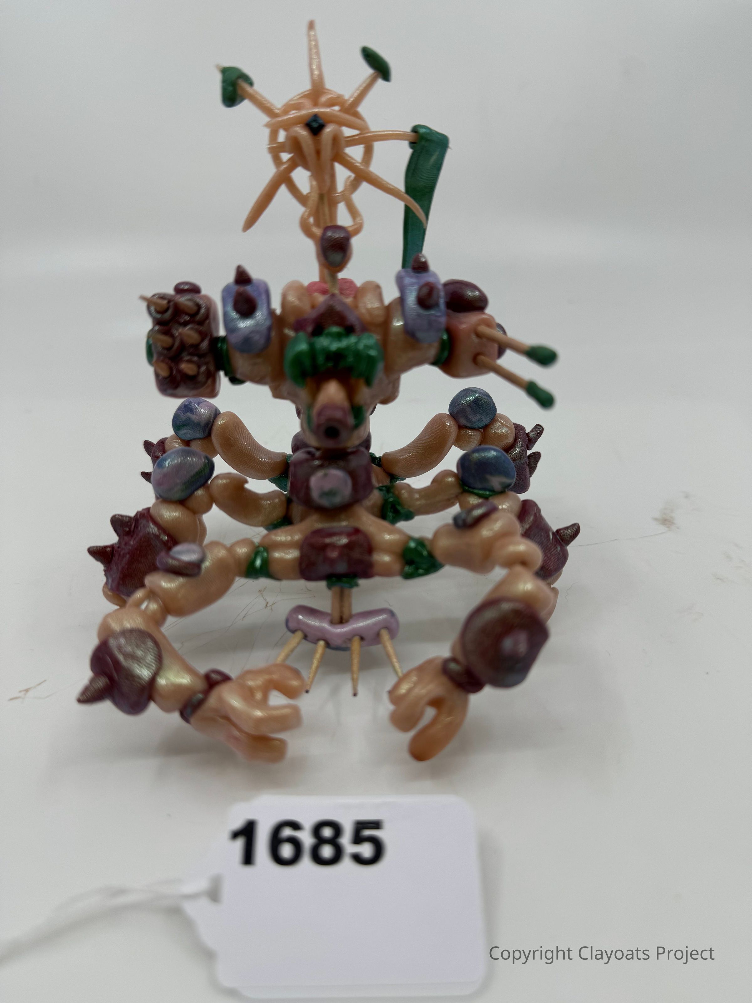 The image displays a unique, abstract clay sculpture with an intricate design composed of interwoven shapes and vibrant colors, giving it a dynamic and fantastical appearance. The piece features various organic forms resembling limbs and spikes, and is embellished with a mix of purple, green, and beige hues. The overall composition showcases the artist's imaginative style and attention to detail, making