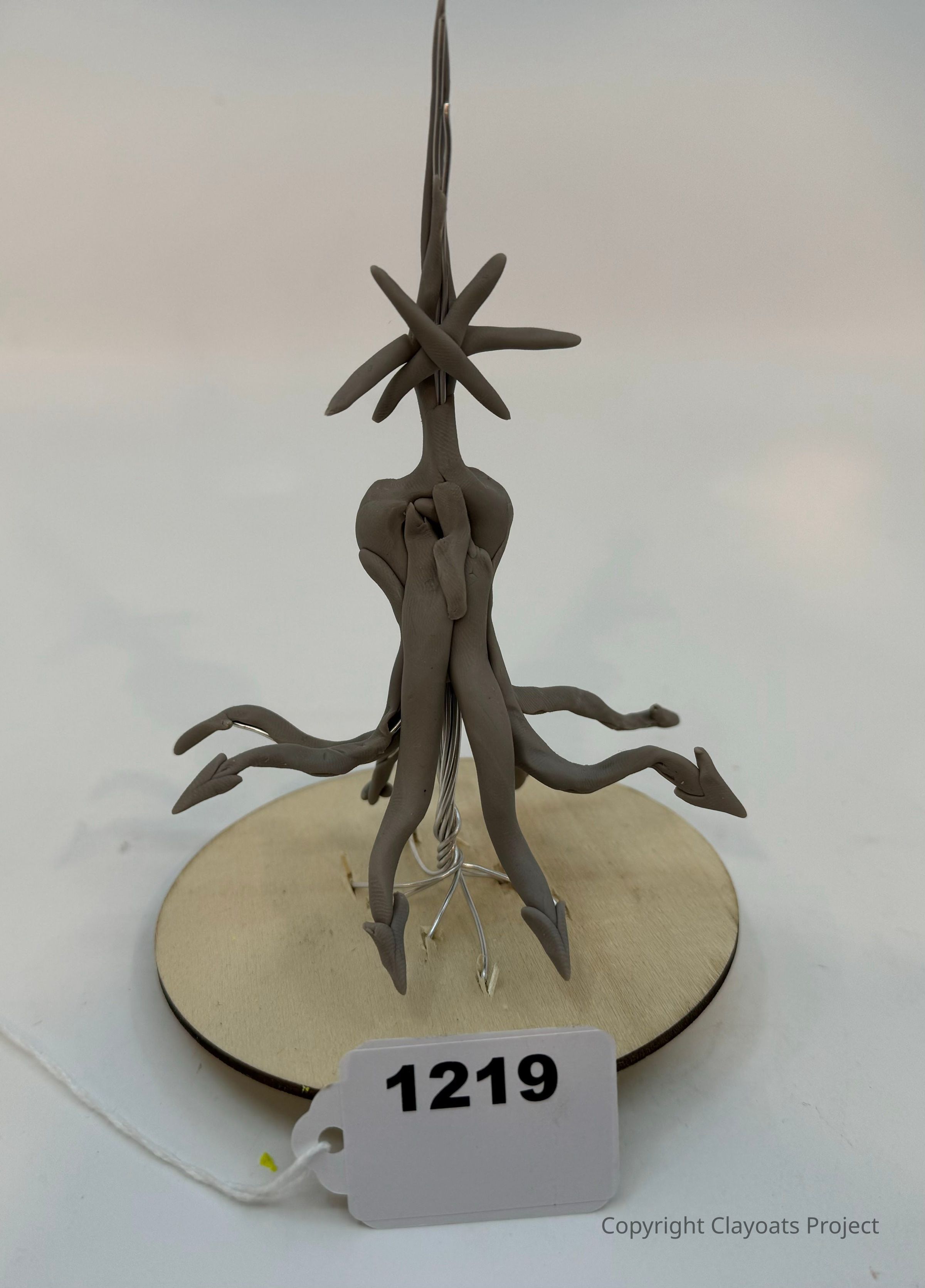 The image shows a unique sculpture made of intertwined clay arms, spiraling upwards in a dynamic form, resembling an abstract, organic structure with elongated limbs extending outward. It is mounted on a circular wooden base,. The sculpture is set against a plain, neutral background, which emphasizes the complexity and creativity of its design. The piece appears to be a conceptual artwork, playing with movement