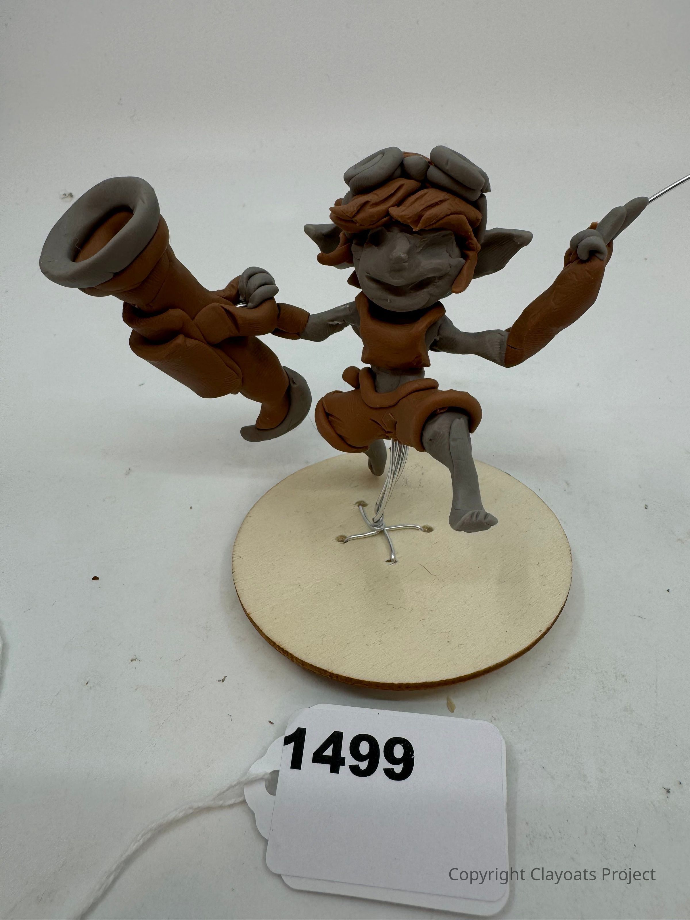 The image showcases a whimsical clay figure of a character with exaggerated features, such as large pointed ears and a wide smile, brandishing a large cannon in one hand while balancing on one foot with an energetic pose. It is perched on a circular wooden base with a metal wire stand for support. The figure, adorned with accessories like goggles and oversized boots, exudes a playful and adventurous personality.
