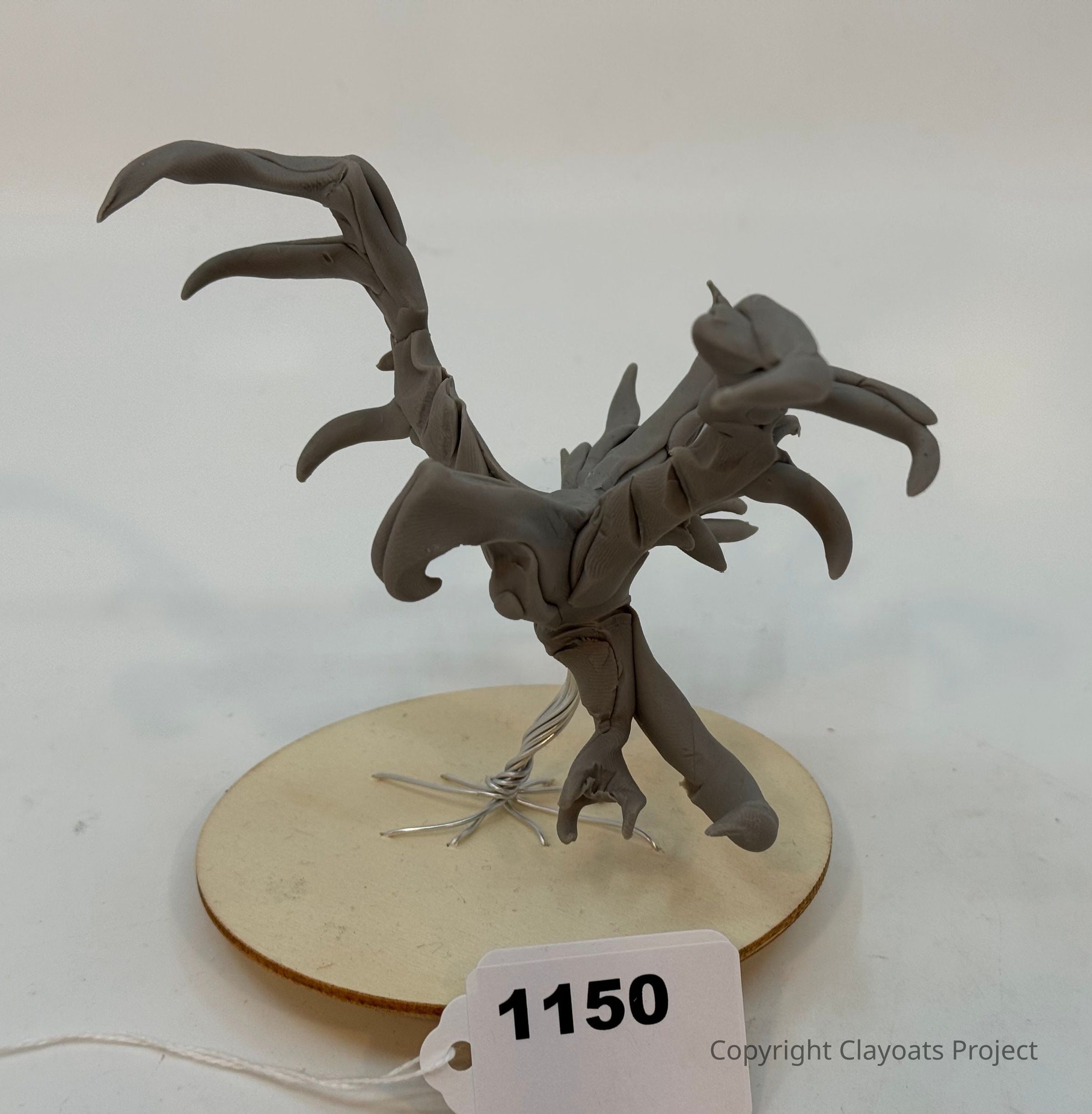 The image features a gray clay sculpture of a whimsical tree on a round wooden base, with curling branches extending outward in various directions, resembling an abstract or fantastical form. The sculpture uses silver wire to suggest roots or a trunk, adding an element of structural support, The background is a plain white surface, allowing the intricate details and creative design of the sculpture to stand out