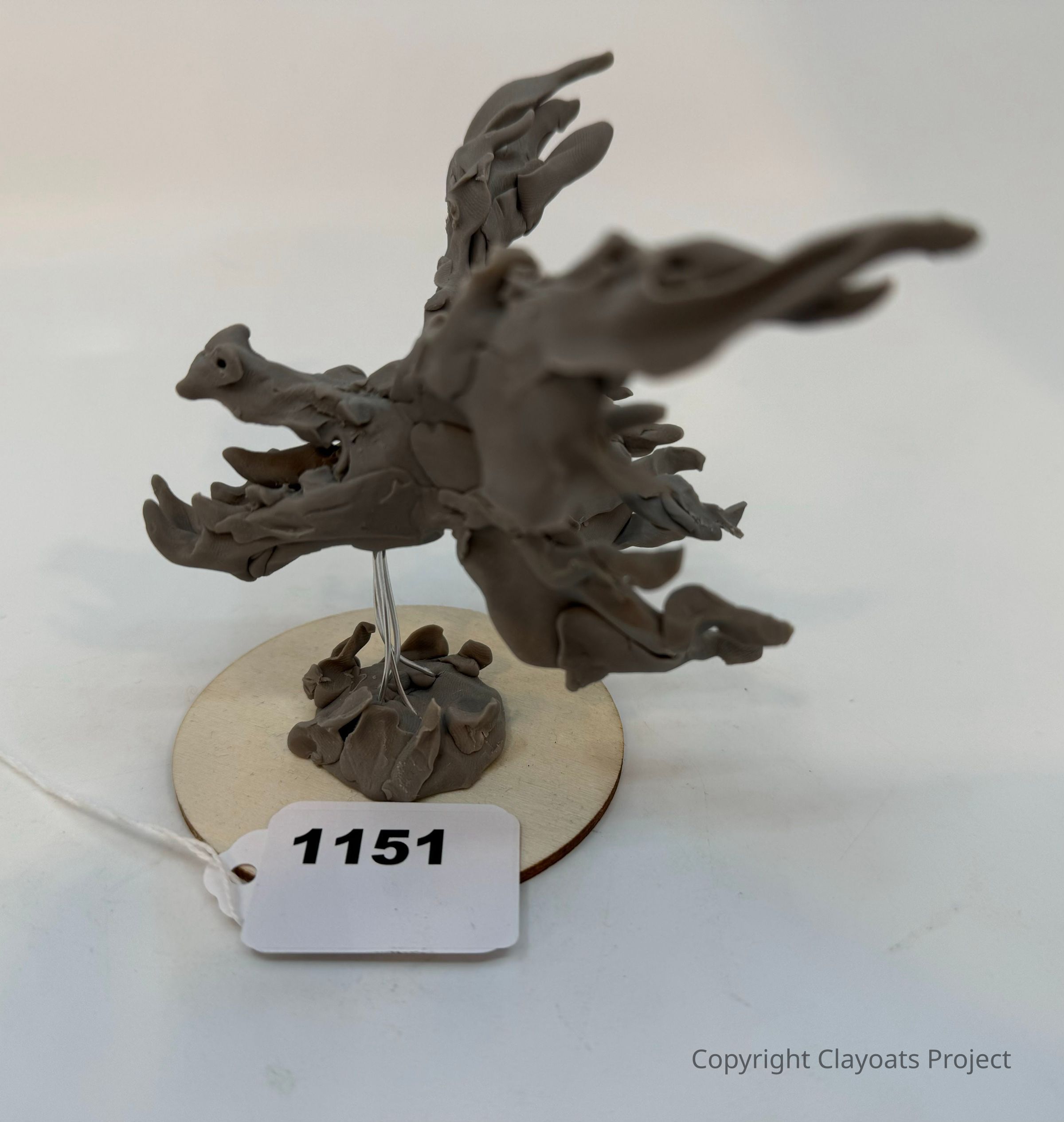 The image shows a clay sculpture on a circular base, featuring abstract, flowing forms that resemble a dynamic, wing-like structure. The sculpture is intricately molded, displaying detailed textures and layers, suggesting motion and fluidity. It rests on a wooden base with a metallic wire frame supporting its elements. The overall impression of the piece is artistic and immersive, inviting viewers to interpret its organic shapes and possible meanings.