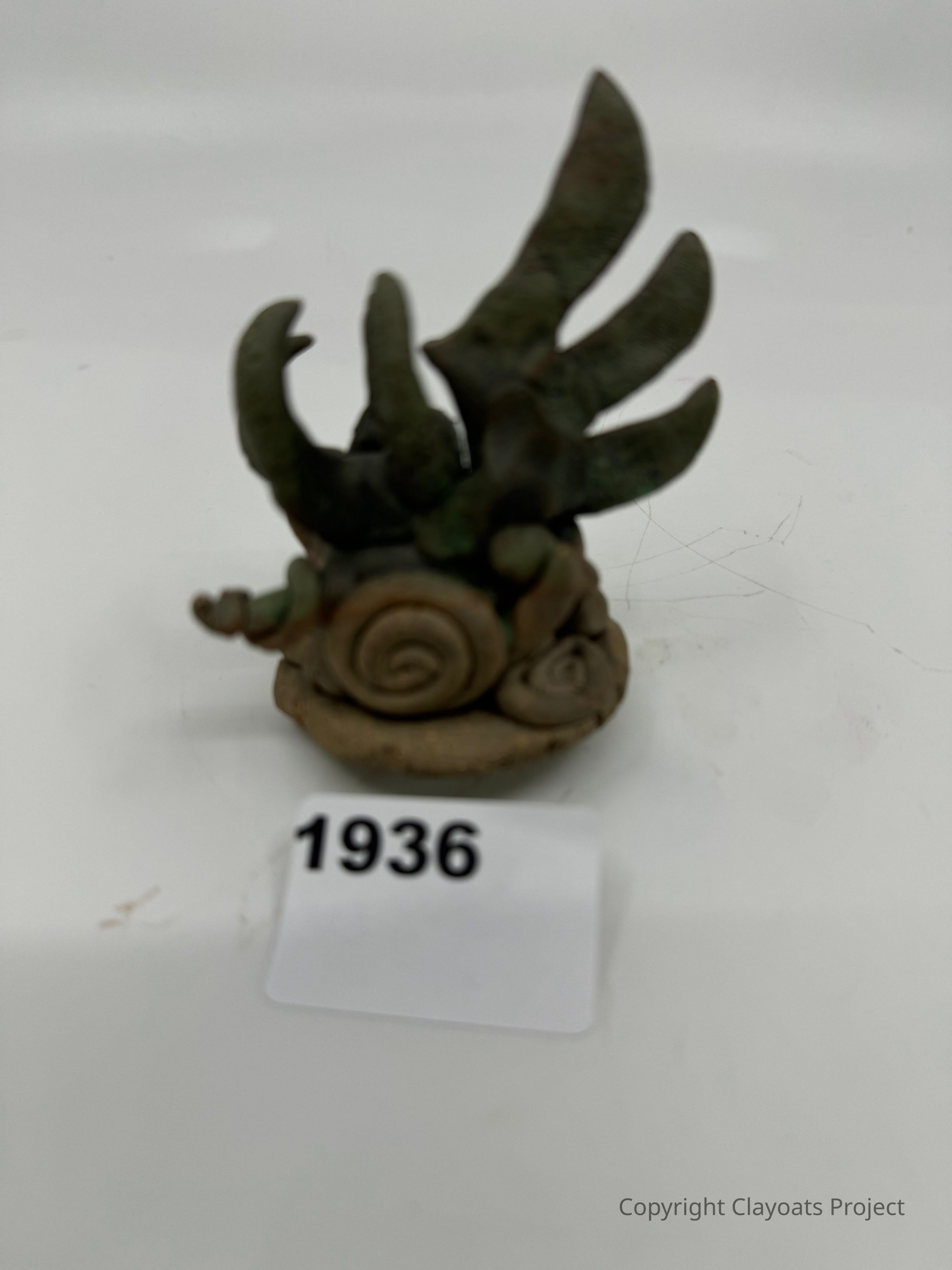 The image depicts a small, sculpted object resembling a snail with exaggerated, ornate features, such as detailed swirling patterns on its shell and large abstract shapes protruding upwards, all in an earthy tone; which sits on a plain light-colored surface in a neutral setting, drawing attention to its intriguing design and artistic craftsmanship, embodying a fusion of natural and imaginative elements, possibly symbolizing creativity and artistic expression in its